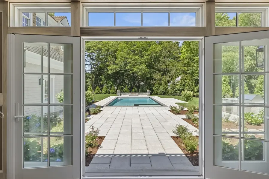 This image captures a breathtaking view through elegant French doors that frame a sophisticated outdoor pool area like a living artwork. The interior perspective showcases beautifully crafted white-painted French doors with traditional multi-pane gla