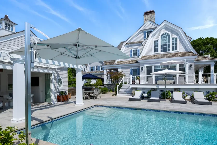 A backyard with a swimming pool, lounge chairs, umbrellas, and a large white house with multiple windows and a porch.