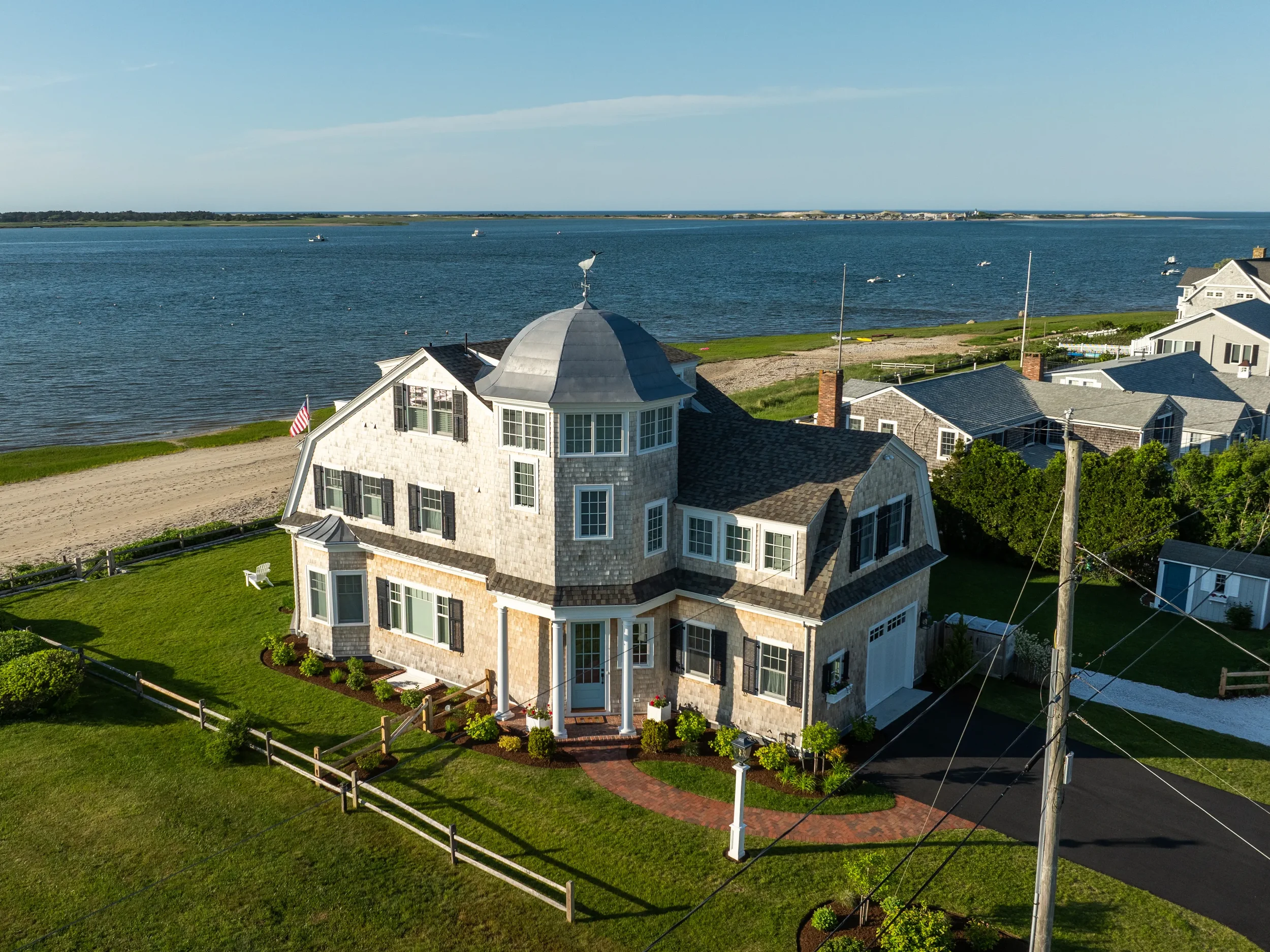 Large beachfront house with a turret, gray shingles, black shutters, and a curved brick walkway leading to the front door. Surrounded by a manicured lawn, overlooking a body of water with boats, and neighboring houses.