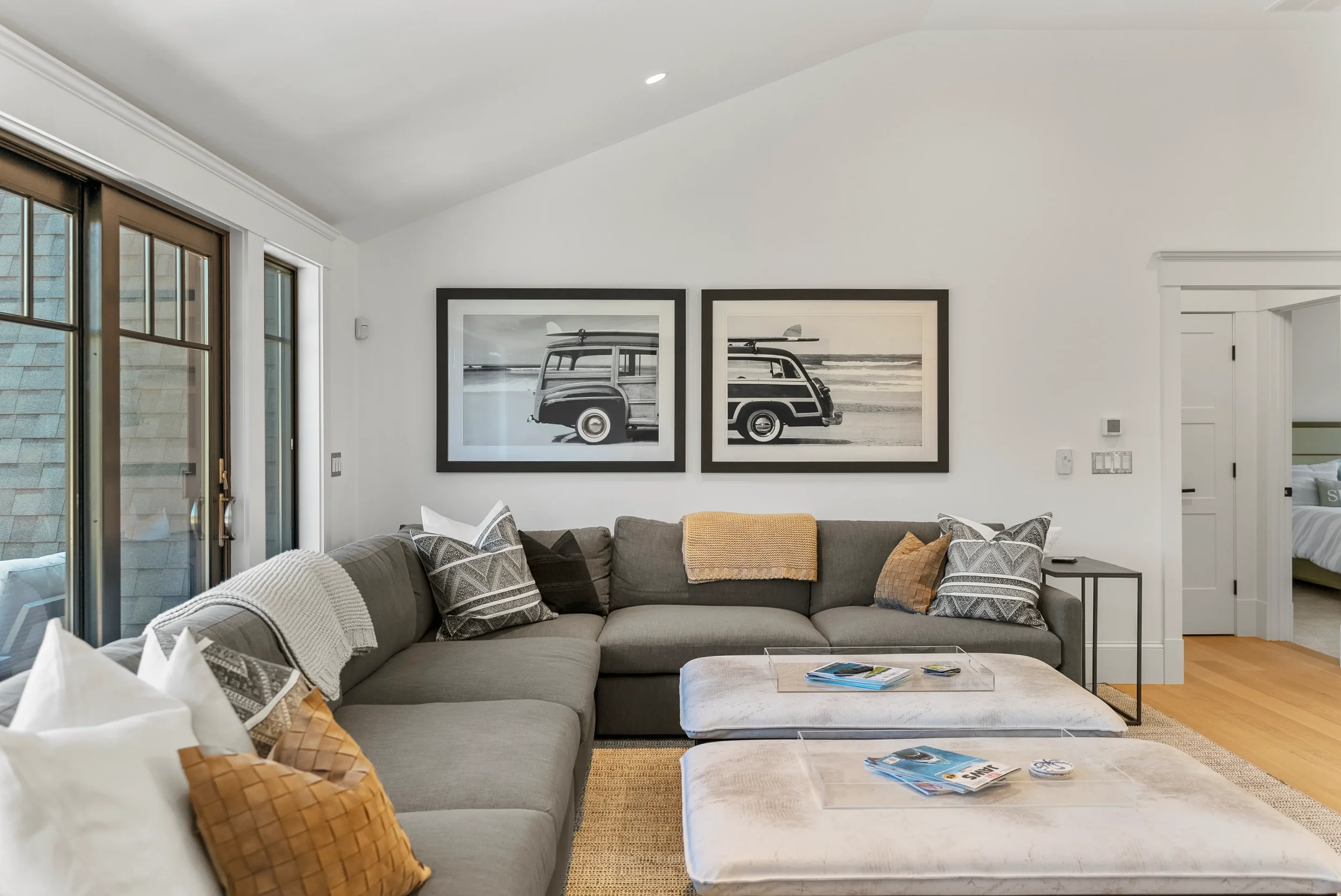 Living room with a gray sectional sofa, decorative pillows, and a throw blanket, two black-and-white framed photographs of a vintage surf van on a beach, sliding glass doors, and a view of a bedroom through an open door.