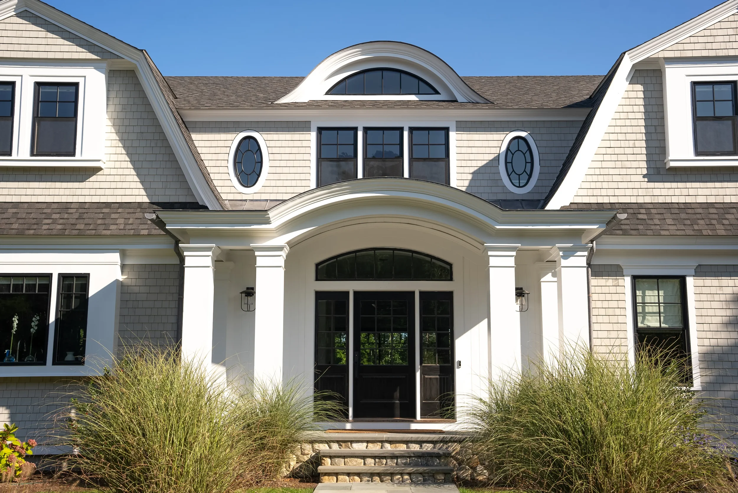 front view of a large, modern house with gray shingles, black windows, white columns, and a curved white trim over the entrance. There are plants with tall grasses in the front yard and a blue sky overhead.