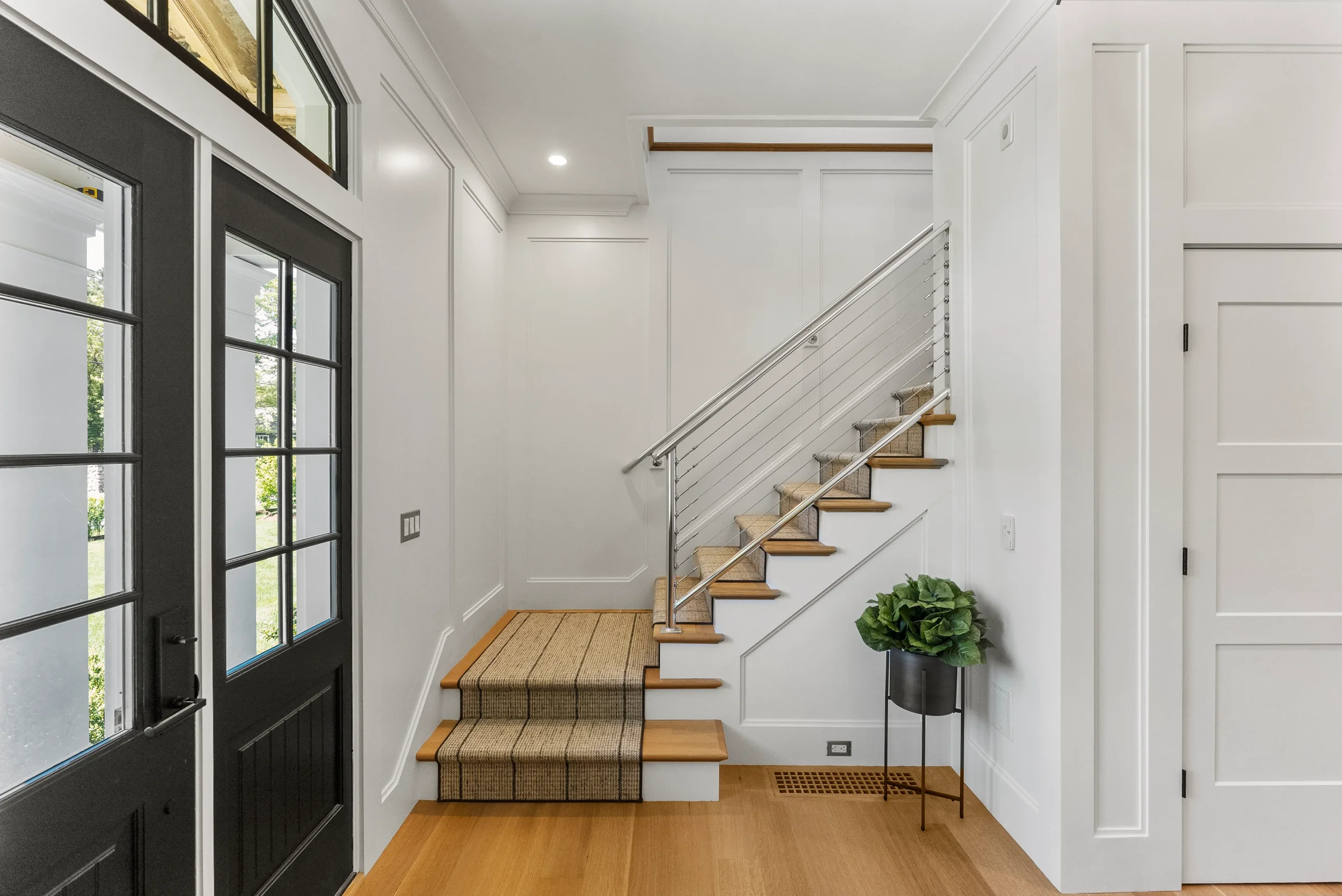 Interior view of a modern home entryway with a staircase, black front door, hardwood flooring, and a potted plant.