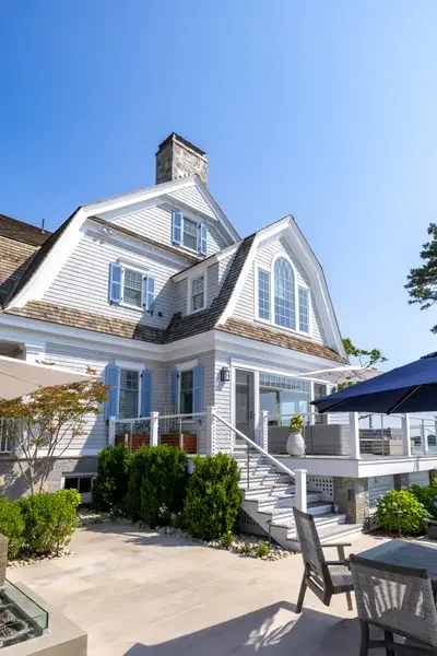 Front view of a large white house with blue shutters, a deck with outdoor furniture, and a blue umbrella, under a clear blue sky.