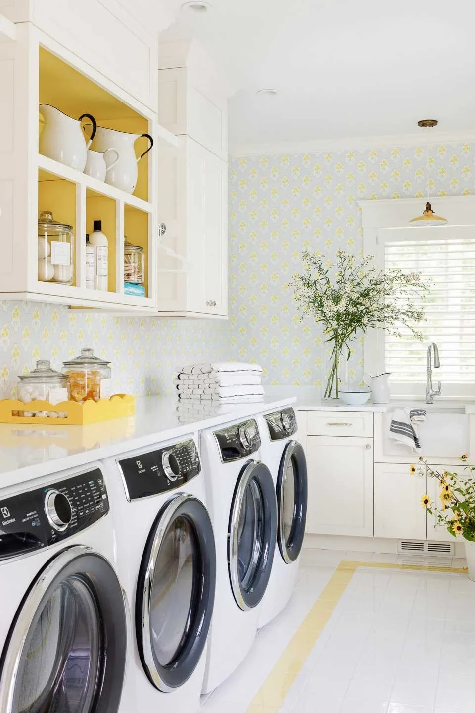 Laundry room with three front-loading washer and dryer, white cabinets, a countertop with towels, glass jars, and decorative dishes, a vase with green branches, and a window with blinds.