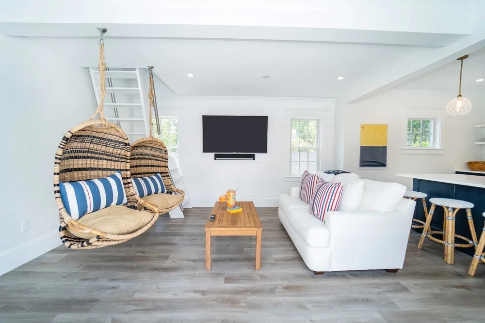 Living room with two hanging woven basket chairs, a white sofa with striped pillows, a wooden coffee table, a wall-mounted TV, and bar stools at a kitchen counter.