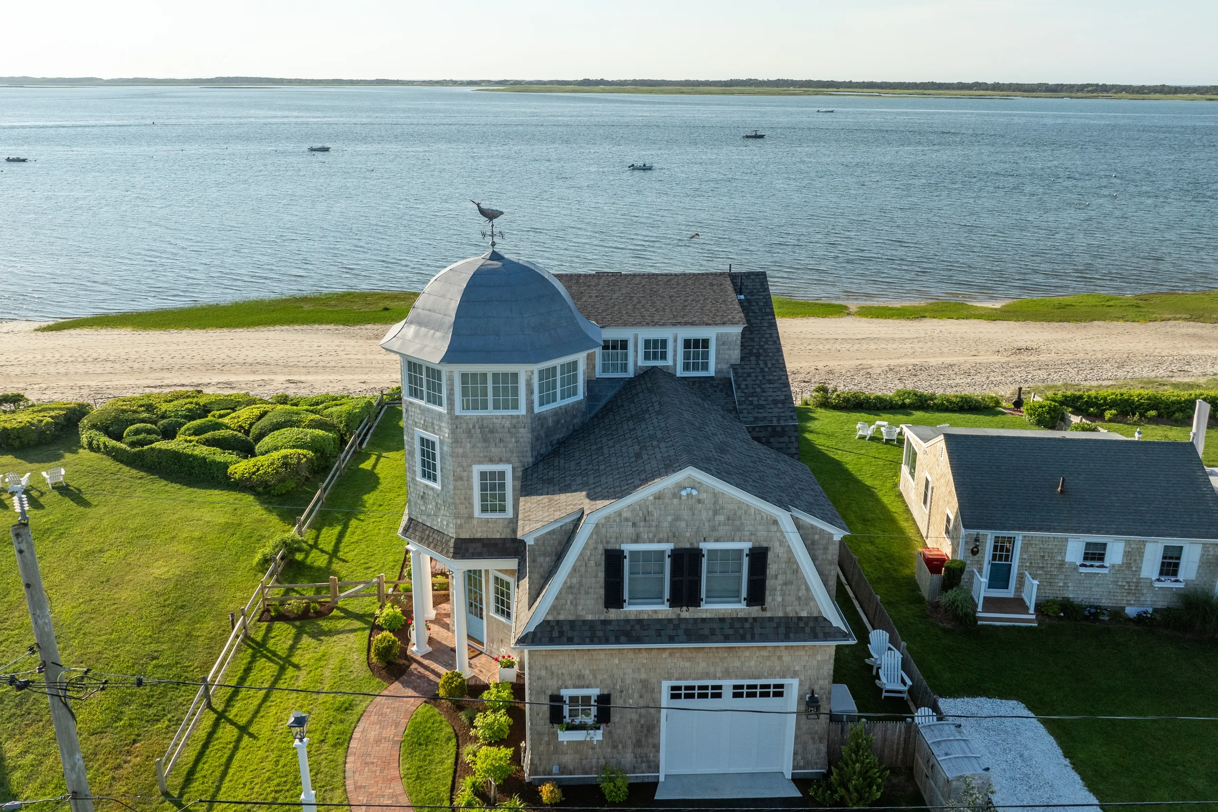 A house by the water with a sandy beach, boats, and green grass in the background, featuring a turret with a weather vane.