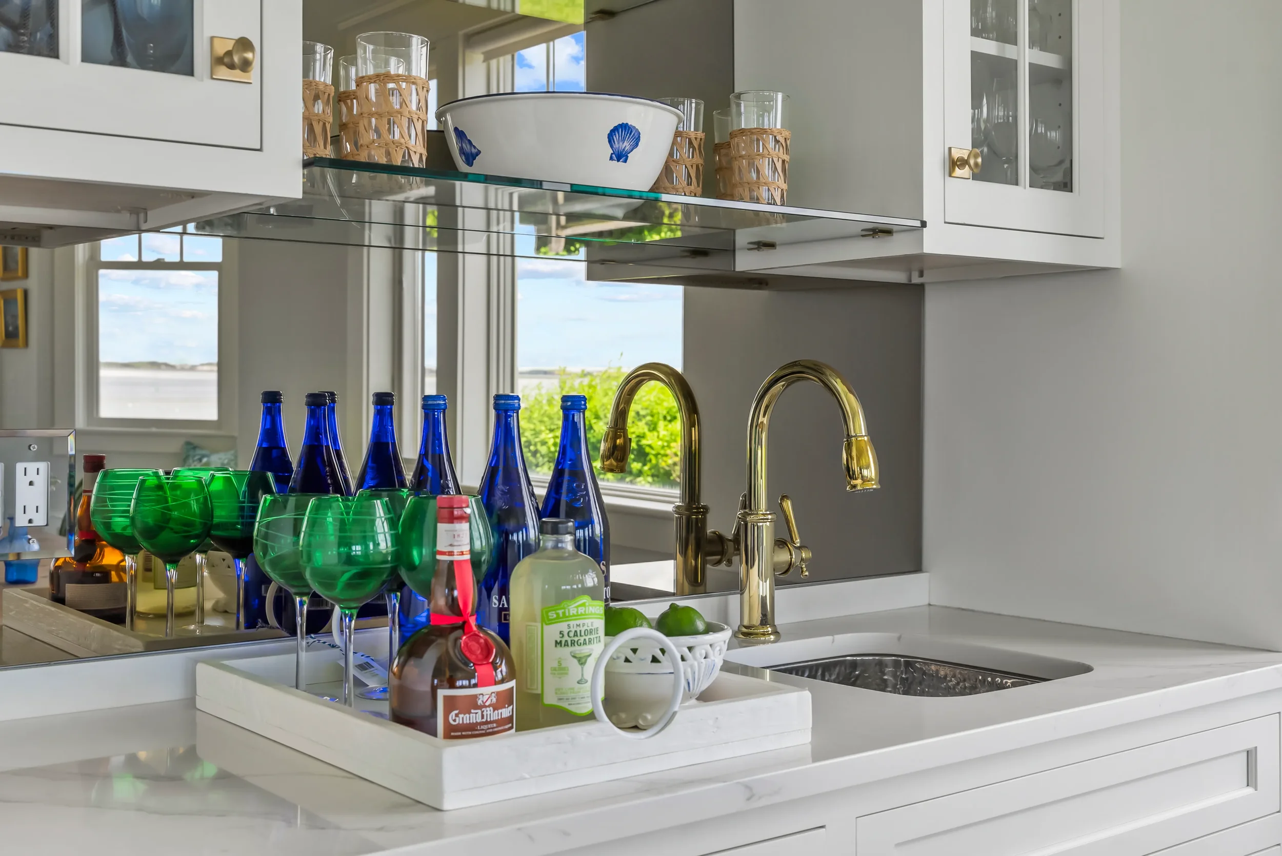 Kitchen sink area with gold faucet, blue bottles, green glasses, and a tray with liquor and lime juice bottles.