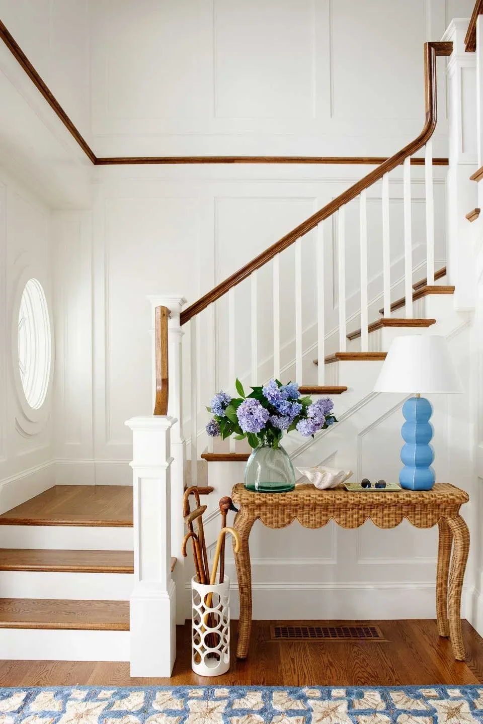 Interior view of a home entryway with a staircase, a wicker table with blue vase and flowers, a lamp, and umbrellas.