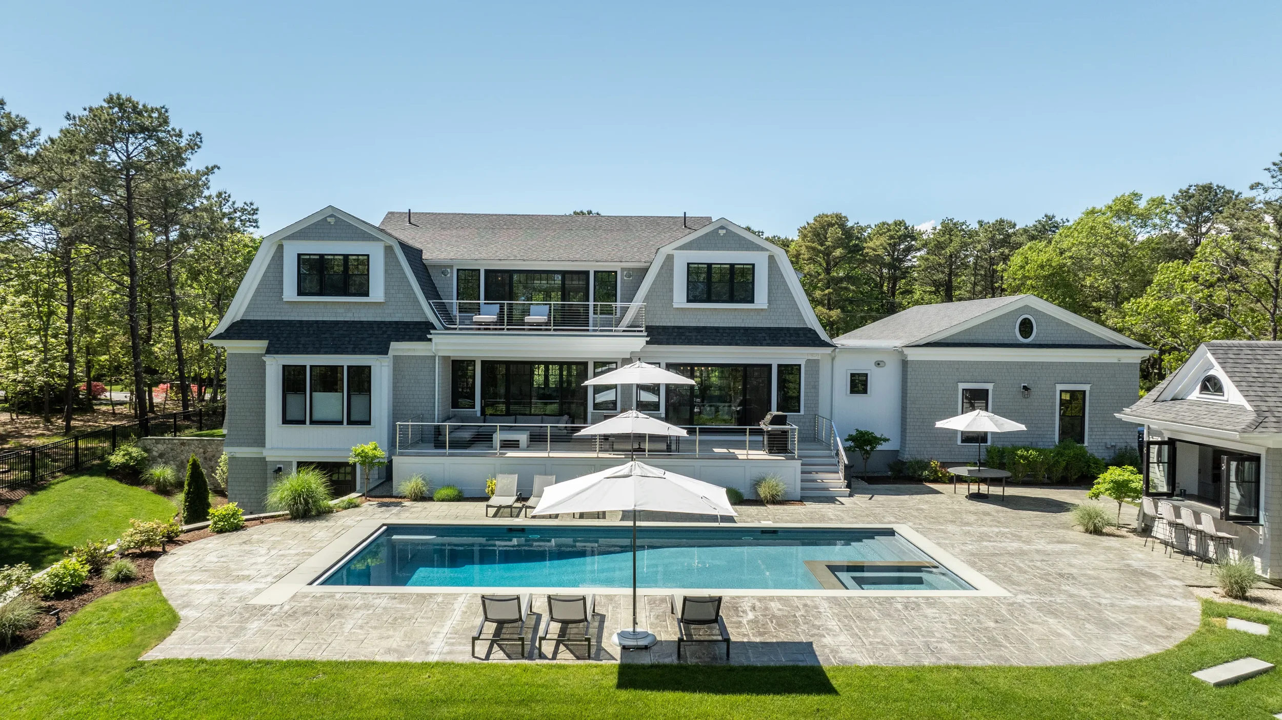 A large modern house with a backyard and swimming pool, surrounded by trees, with outdoor furniture and umbrellas.
