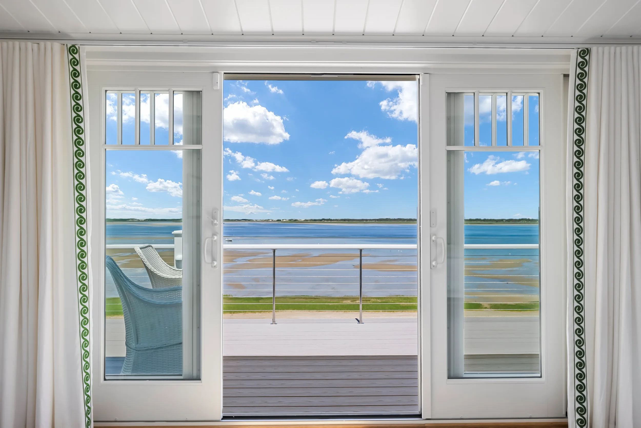 View of a balcony overlooking a body of water, seen through sliding glass doors, with white curtains on either side, green trim, and a blue sky with clouds.