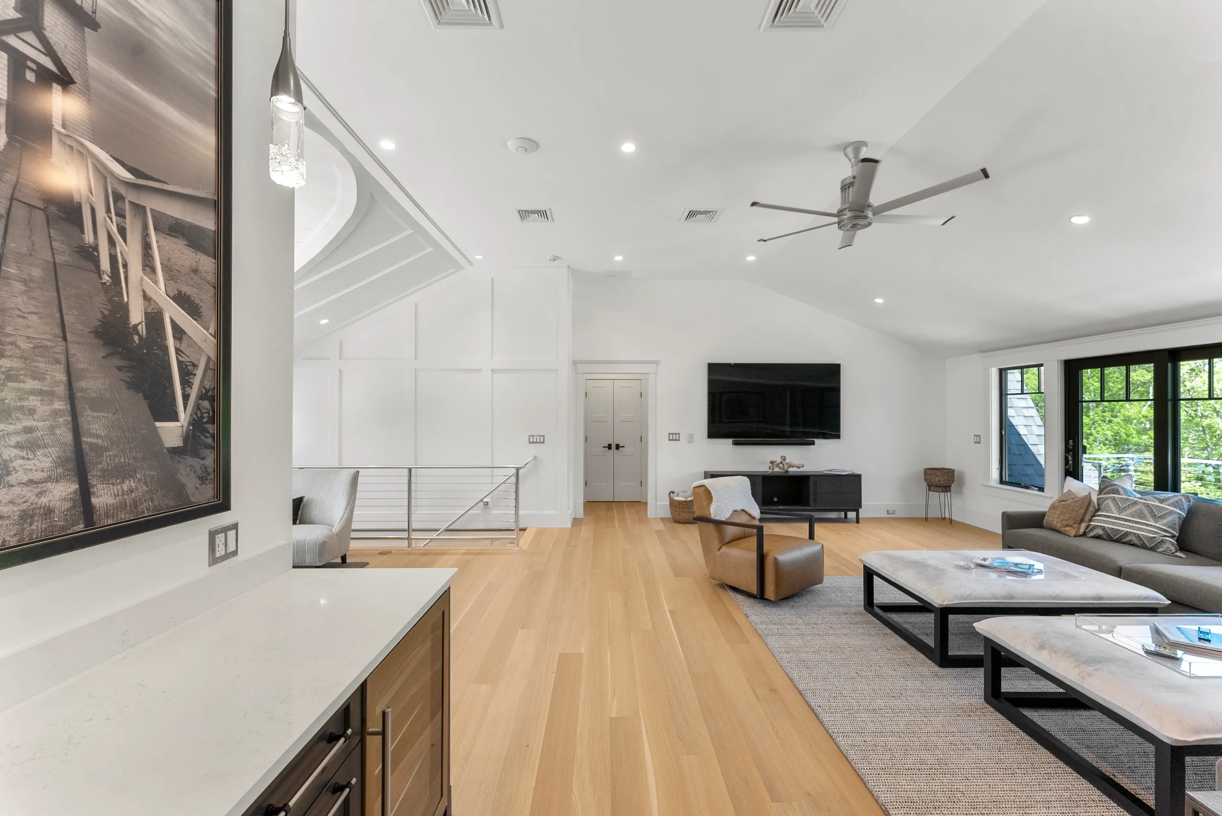 Spacious modern living room with white walls, hardwood floors, a wall-mounted television, and large windows with outside views. Furniture includes a gray sofa, brown armchair, and coffee tables, with a ceiling fan and ceiling lights.