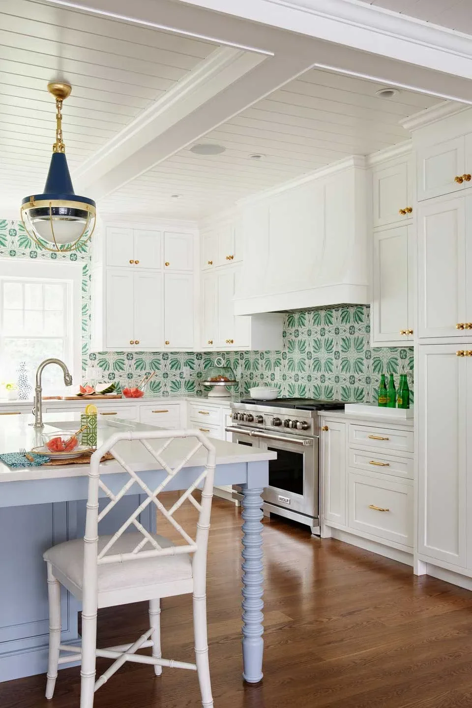 This image showcases a bright, inviting kitchen interior that blends traditional charm with modern flair. The cabinetry is crisp white with elegant gold handles, framing a stainless steel gas stove and oven beneath a sleek white range hood. A standou