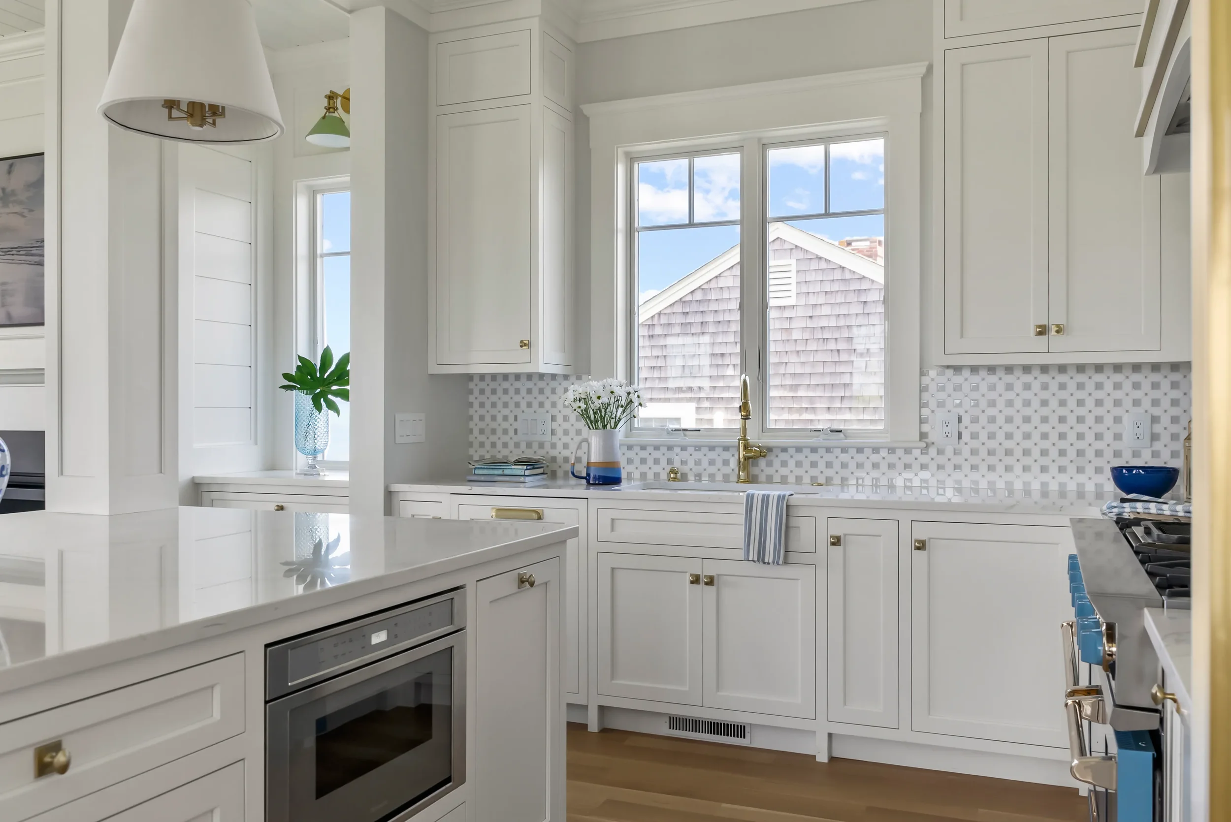 Bright white kitchen with white cabinets, a window above the sink, gold fixtures, a blue bowl on the countertop, a stove with blue knobs, and a view of neighboring houses.