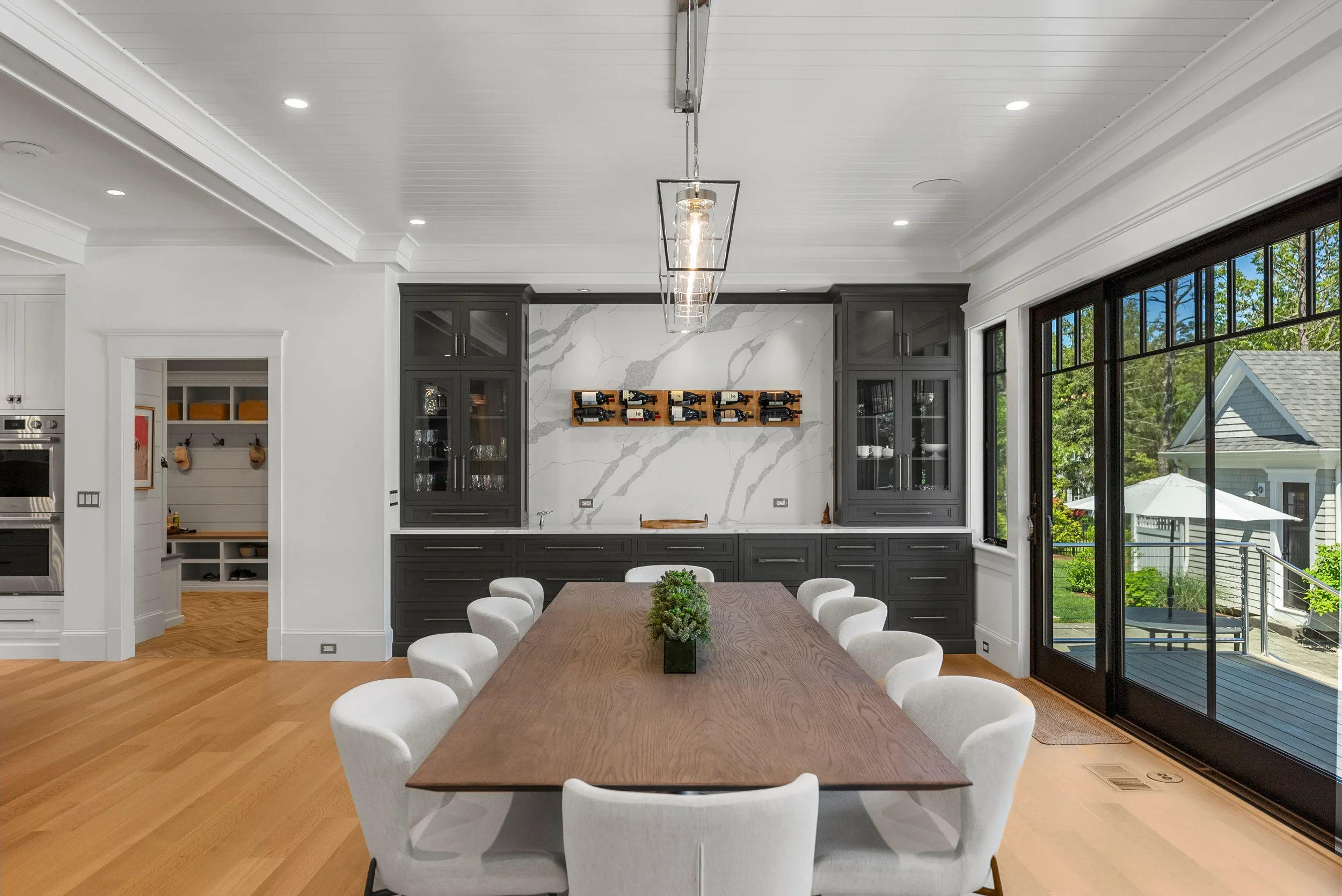Modern dining room with a wooden table, white upholstered chairs, black cabinetry, and large windows overlooking a backyard with a deck and patio furniture.