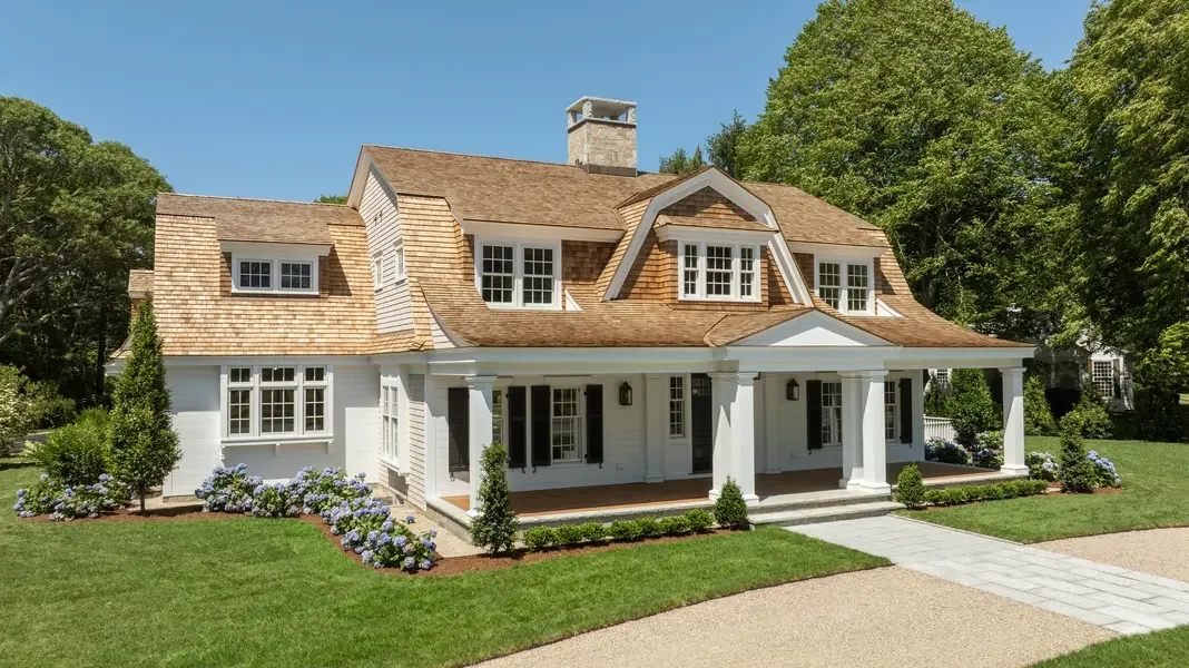 This image showcases a stunning Cape Cod-style estate home set against a brilliant blue sky. The expansive residence features classic New England architecture with weathered cedar shingle siding in natural tones and white trim throughout. The complex