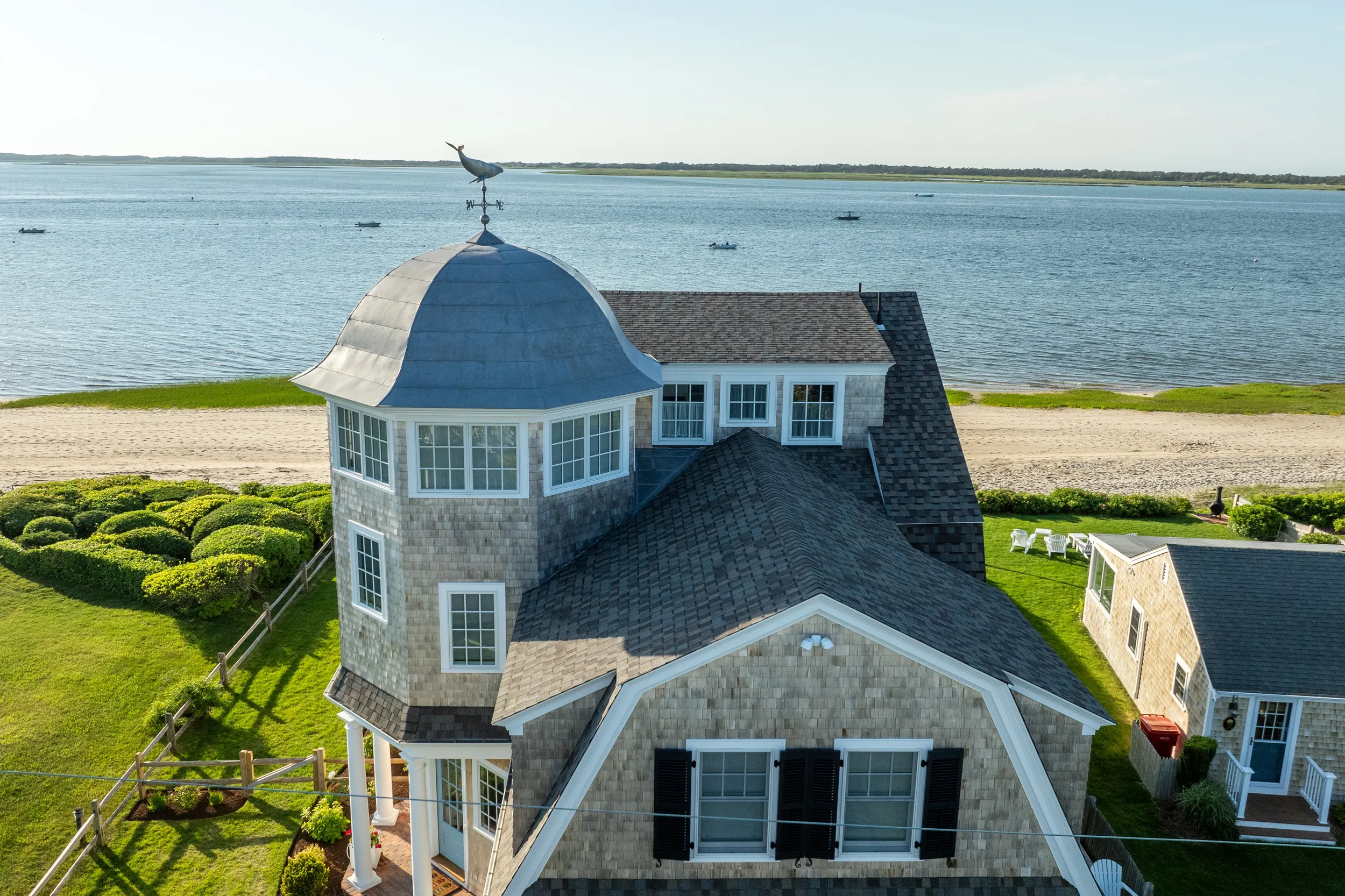 A large house by the water with a turret that has a bird weather vane on top, surrounded by a well-maintained yard.