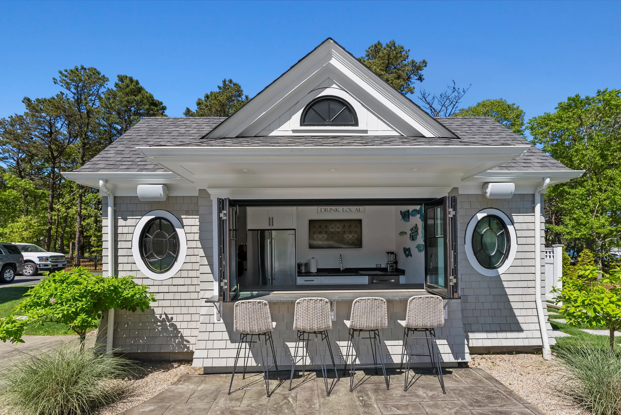 Small house with an open kitchen serving as an outdoor bar, featuring four barstools, a refrigerator, and wall art, surrounded by greenery and parked cars under a clear blue sky.