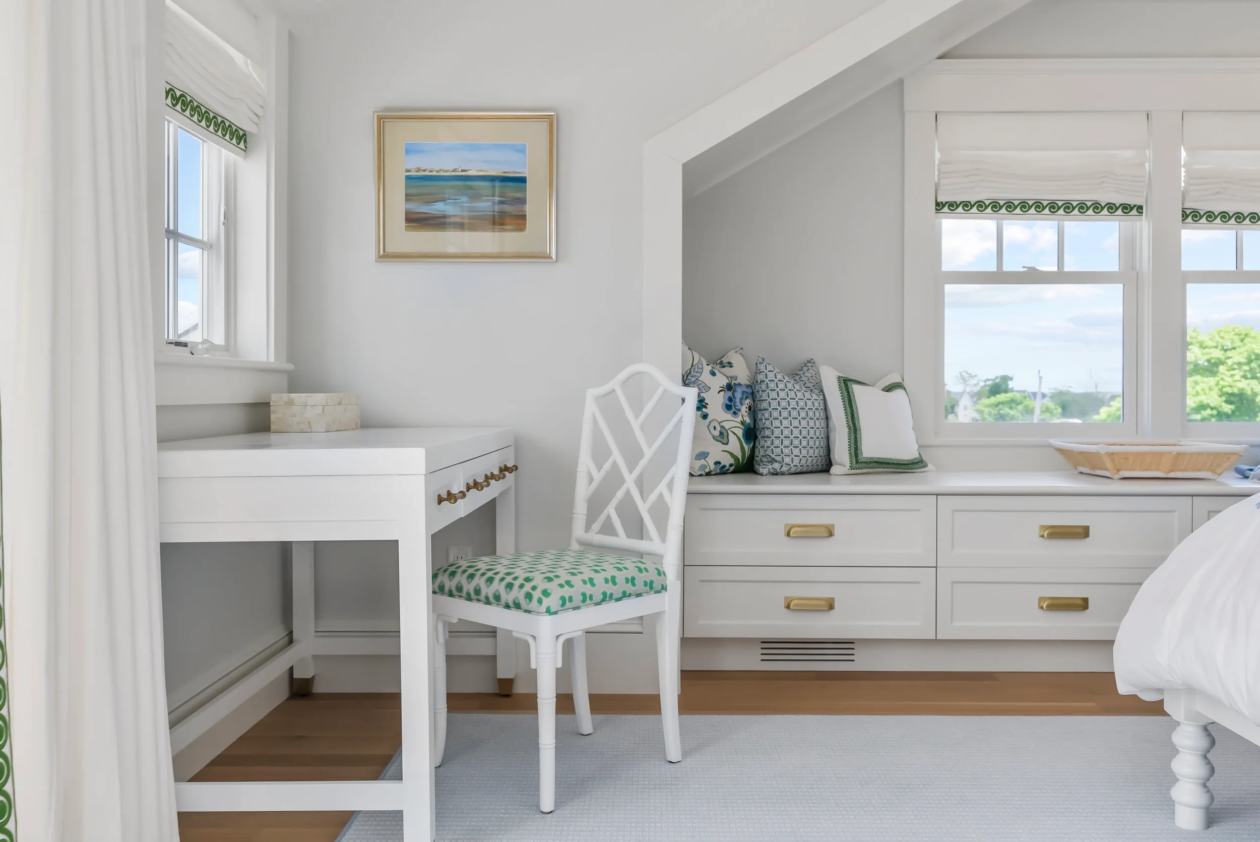 A bright bedroom corner with white walls, a window with white curtains and green trim, a white desk with a patterned cushion, a white chair, and a window seat with multiple pillows under large windows showing green trees and blue sky.