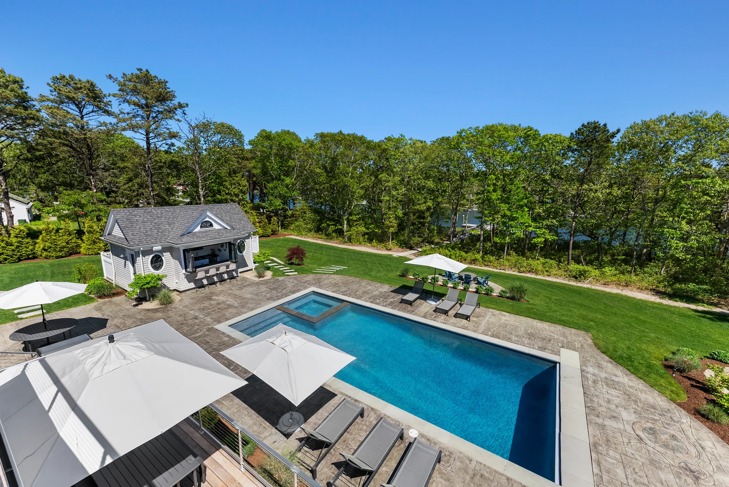 A backyard with a swimming pool, surrounded by lounging chairs, umbrellas, and a small pool house, with trees in the background under a clear blue sky.