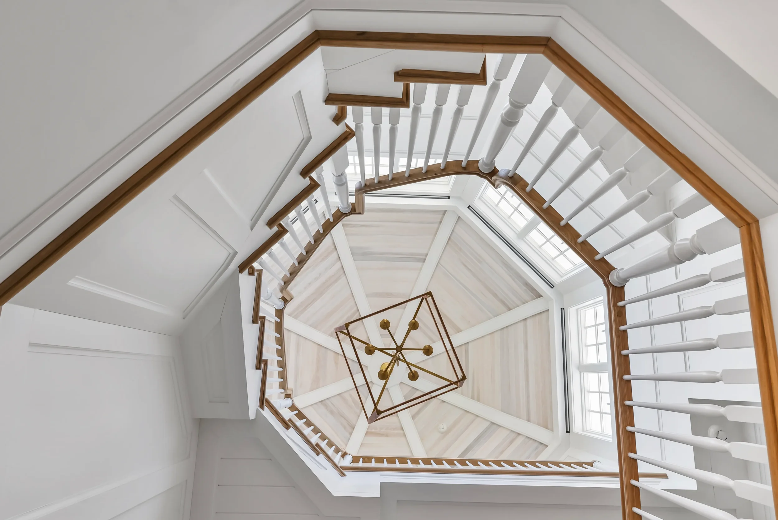 Looking up at a multi-story staircase with white balusters, wooden handrails, and a central geometric chandelier, with natural light coming from large windows.