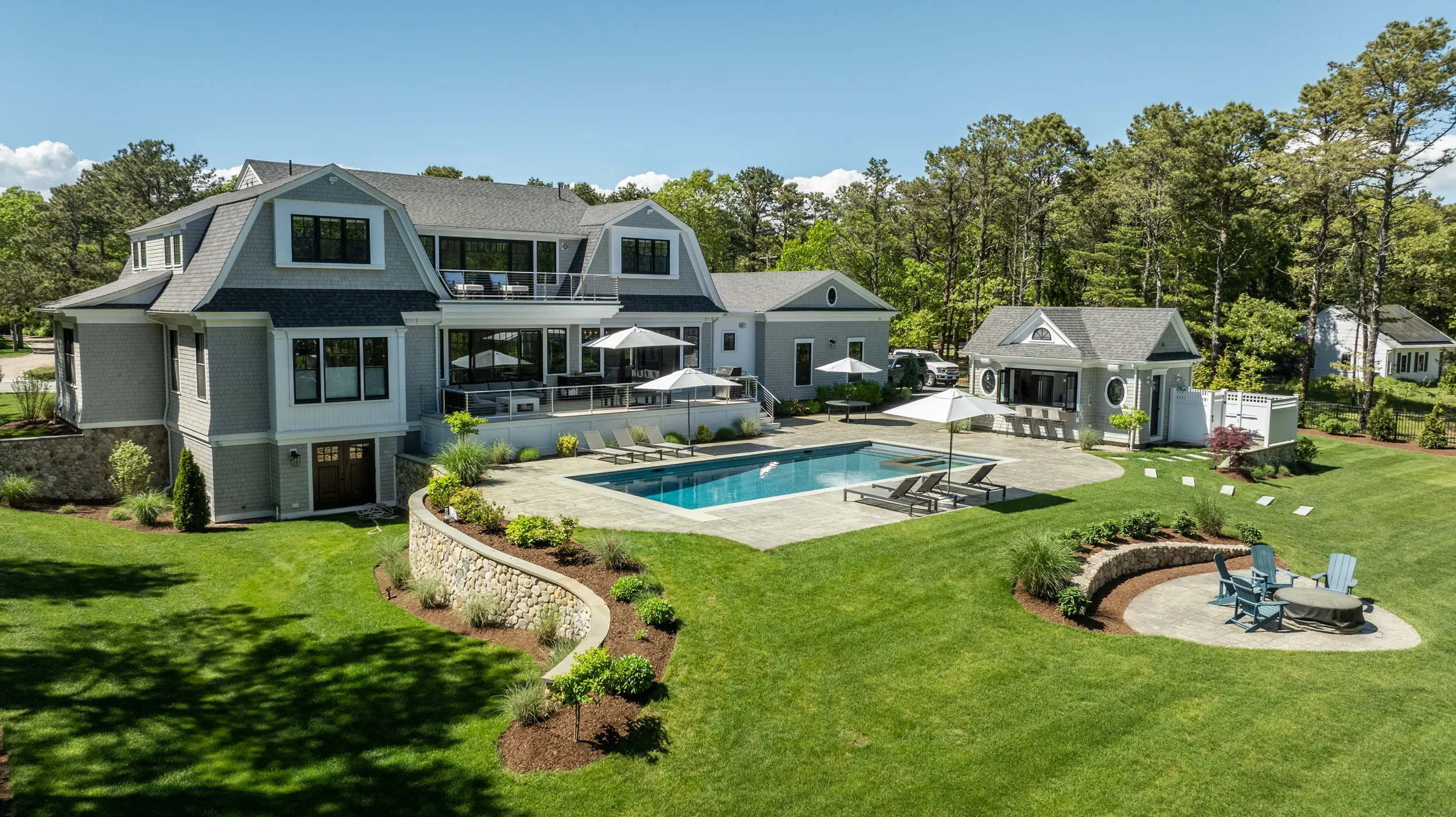A large, elegant house with a backyard pool area, surrounded by lush green grass, trees, and landscaped garden beds, with clear blue skies overhead.