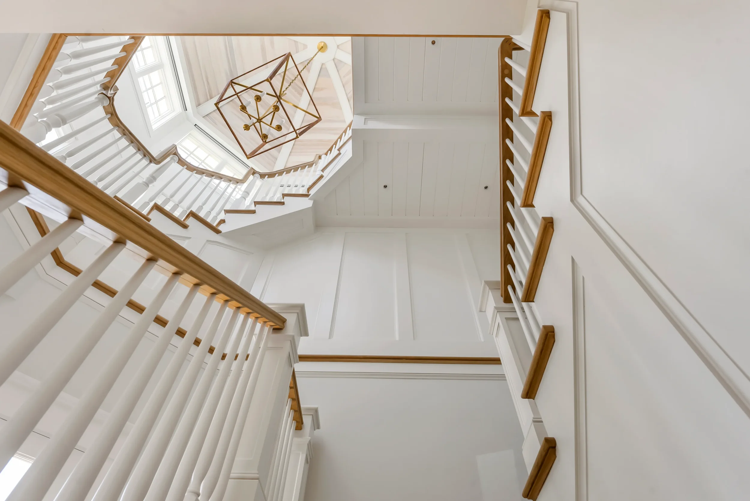 Looking up at a white staircase with wooden handrails and balusters, and a geometric chandelier hanging from the ceiling.