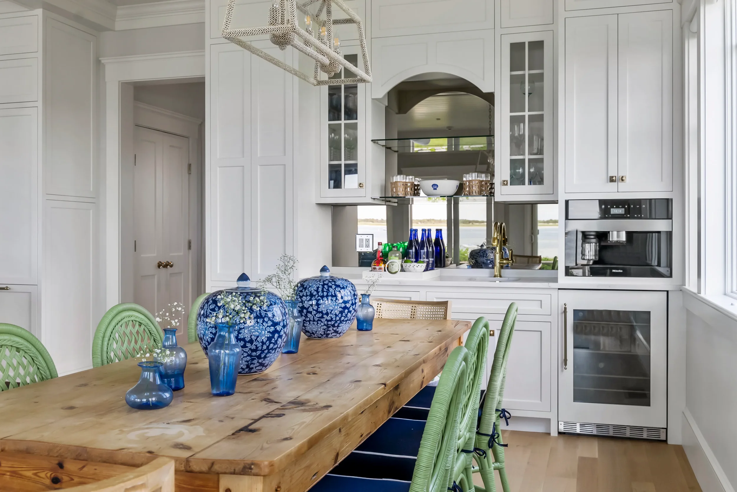 A bright kitchen with white cabinets, a wooden dining table with green chairs, blue vases with flowers, and a view of a lake through large windows.
