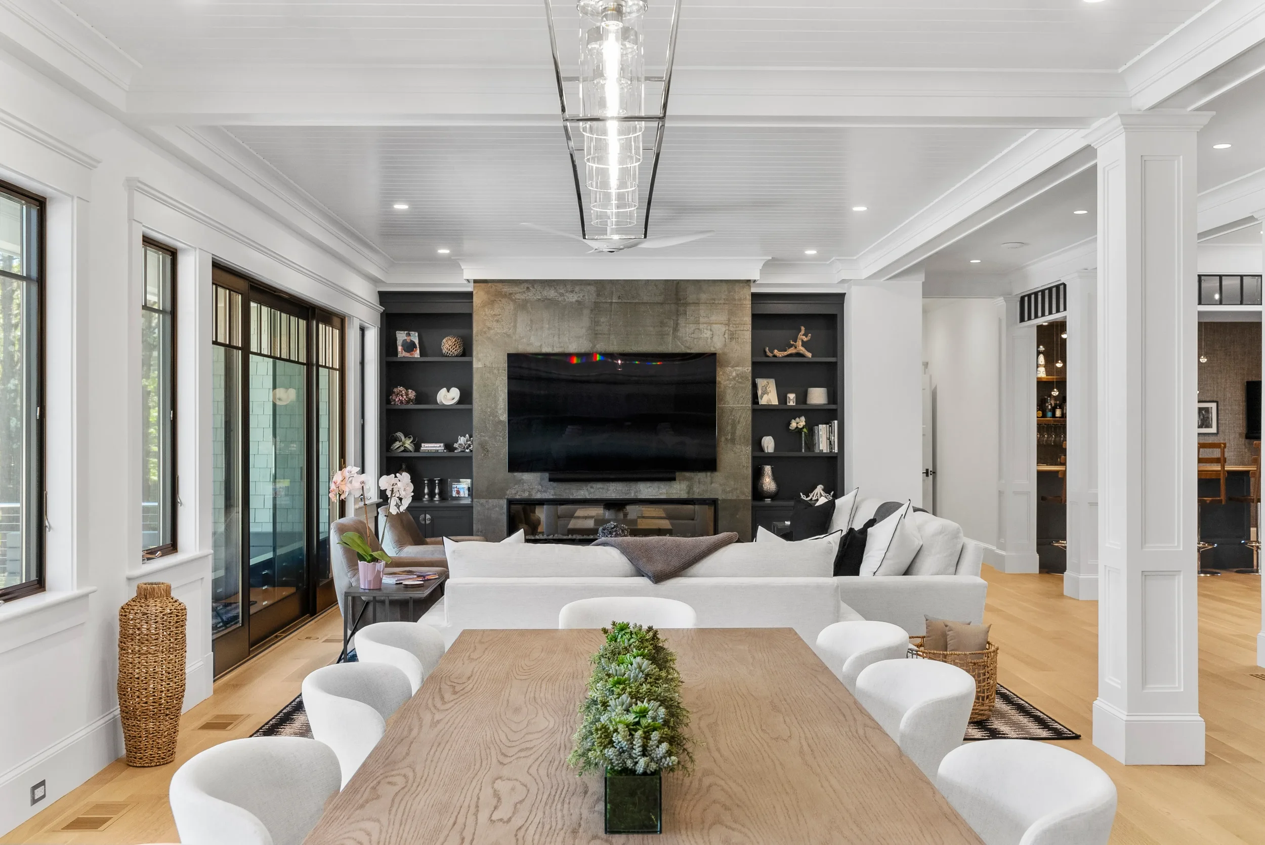Modern living room with white walls, a large flat-screen TV above a fireplace, and built-in black shelves with decorative items. There is a round dining table with a centerpiece of plants, and a white sectional sofa facing the TV. Large windows let i