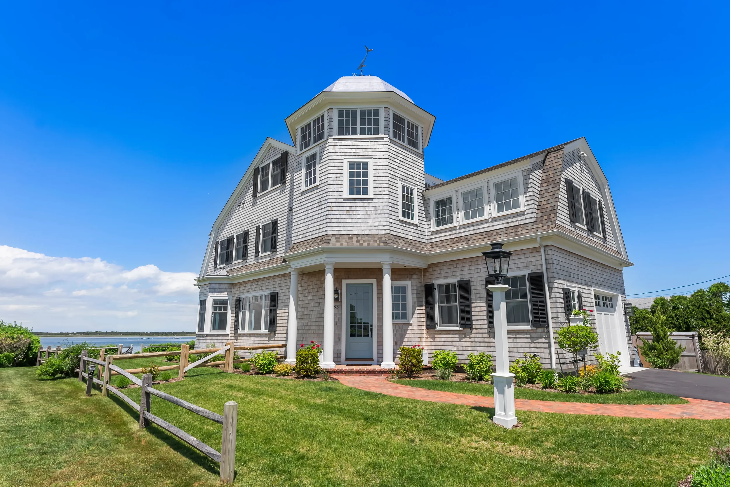 A large, multi-story home with white shingle siding and black shutters, situated on a well-maintained grassy lawn with a brick walkway, a white fence, and a waterfront view under a bright blue sky.