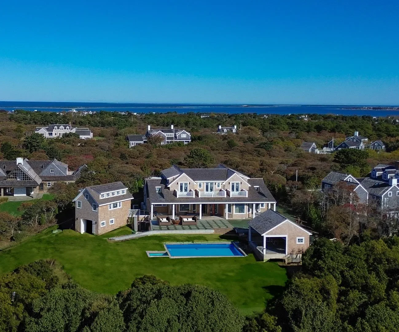 A large house with a backyard swimming pool, surrounded by other houses and trees, with the ocean in the background under a clear blue sky.