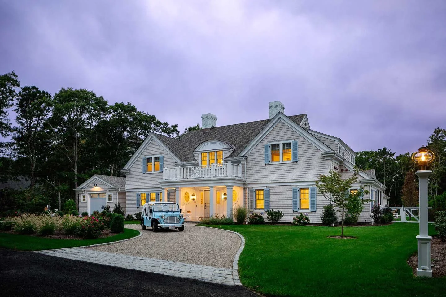 This captivating twilight photograph showcases the same stunning Cape Cod estate bathed in the magical glow of evening light. The home's weathered cedar shingle exterior appears soft and silvery against the dramatic purple-tinged sky, while warm gold