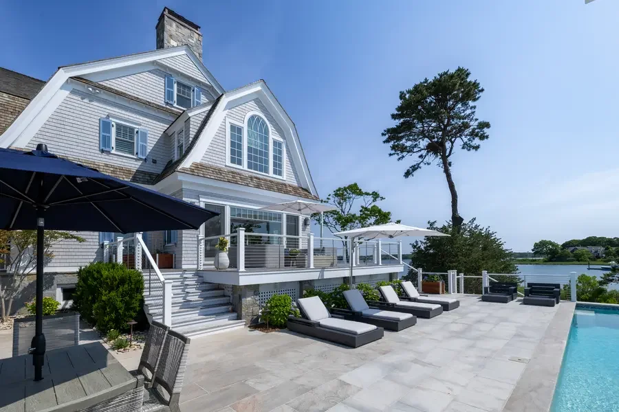 Lakeside house with outdoor pool, lounge chairs, umbrellas, and patio furniture on a sunny day.