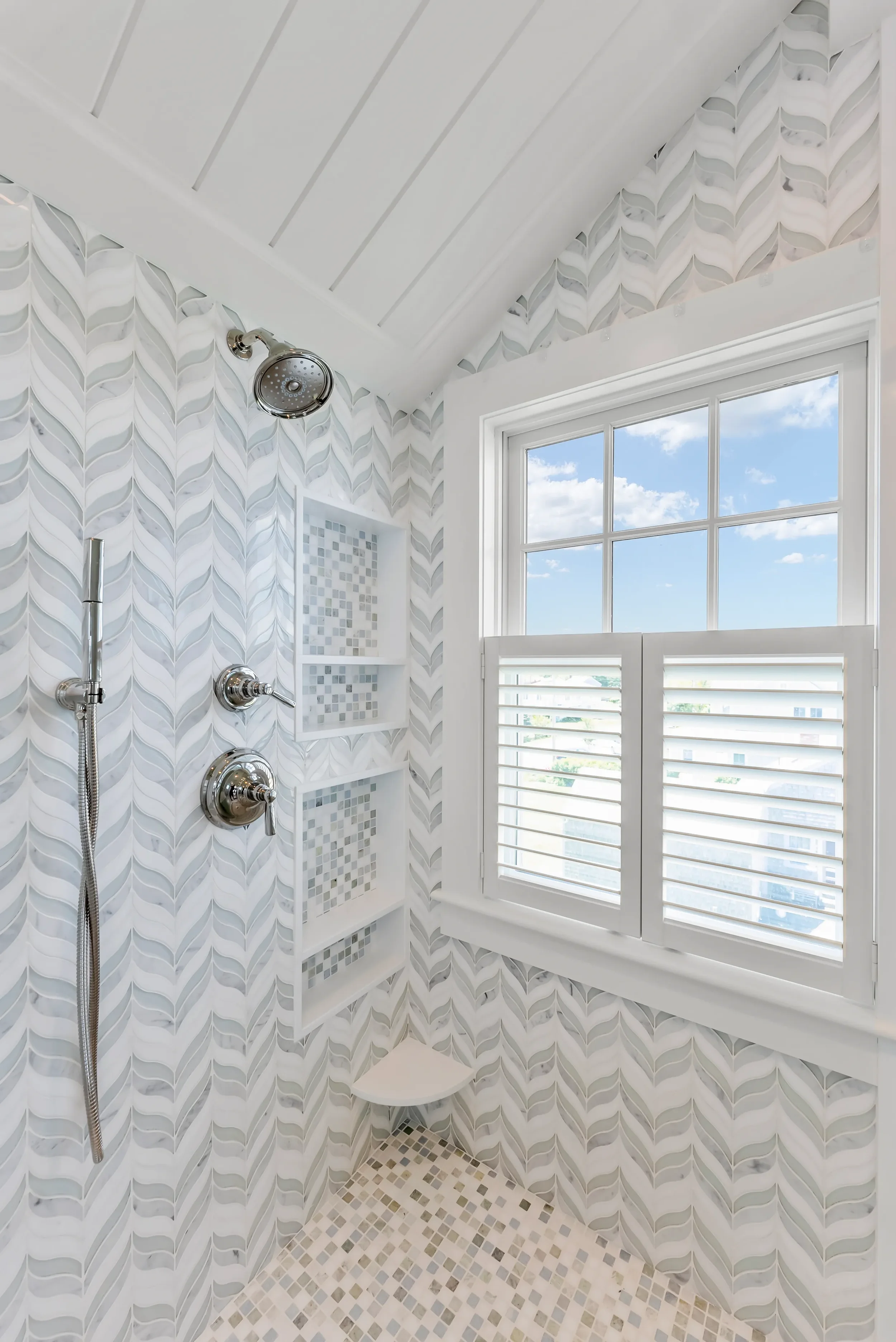 Shower with chrome fixtures, mosaic tile shelves, patterned wall tiles, a window with white shutters, and a small corner shower seat.