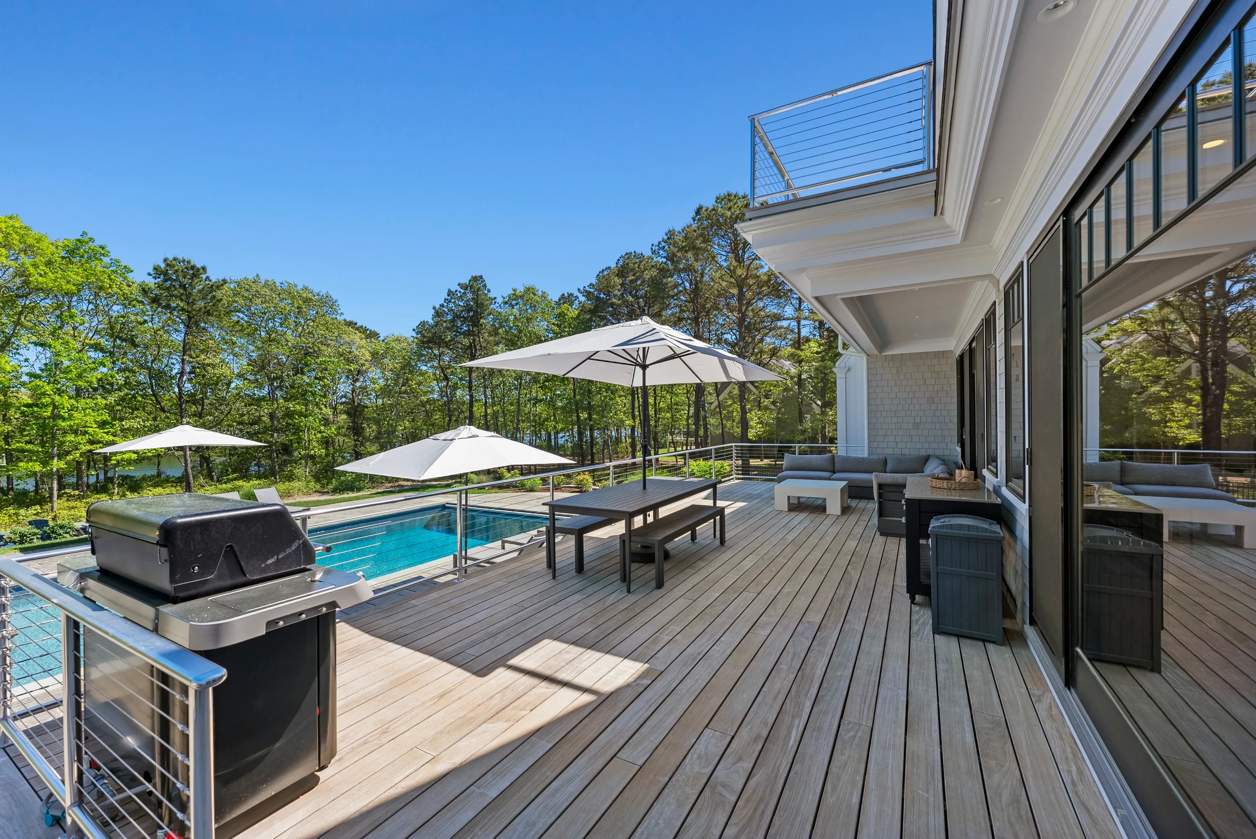 Outdoor deck with a swimming pool, patio furniture, umbrellas, grill, and scenic wooded background.