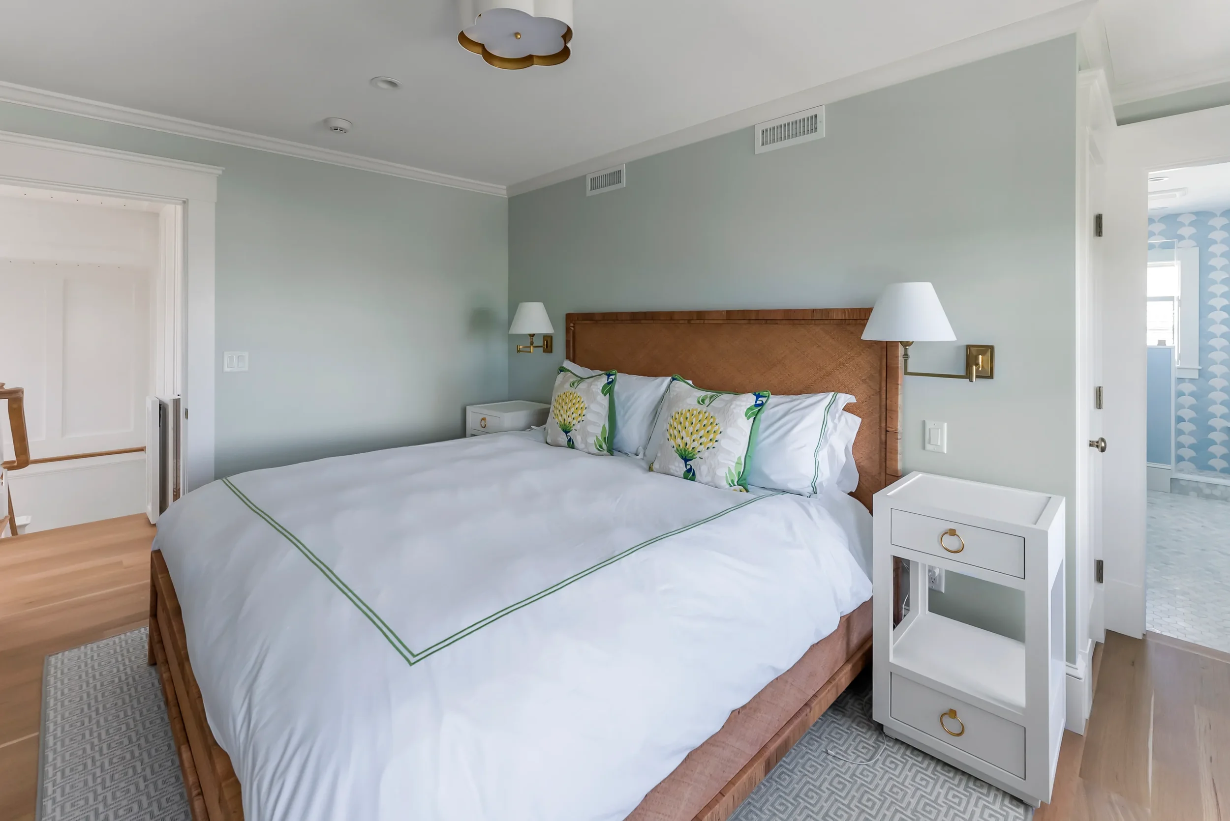 A neatly made bed with white linen and three decorative pillows with green and yellow floral patterns in a modern bedroom with light gray walls, wooden flooring, and white furniture.