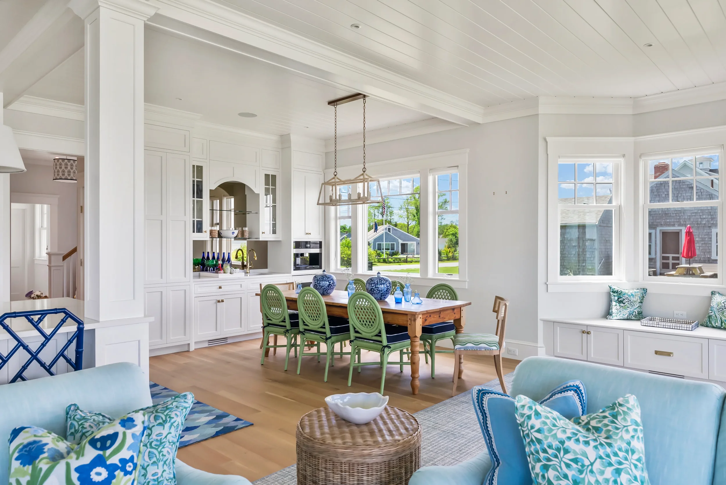 Bright, coastal-style living room and dining area with white walls, light wood floors, and large windows showing a neighborhood scene outside. The space features a white built-in cabinetry, a wooden dining table with green chairs, and a seating area