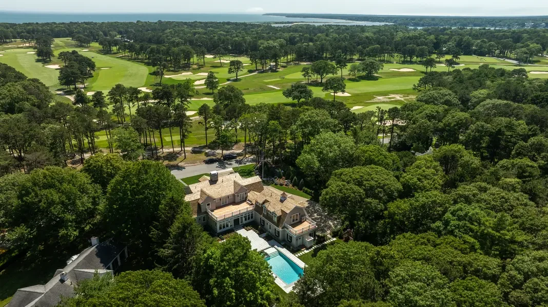 This aerial photograph captures a luxurious estate home nestled within a golf course community. The large, multi-level residence features a distinctive clay tile roof and expansive architecture with multiple wings and levels. A pristine swimming pool