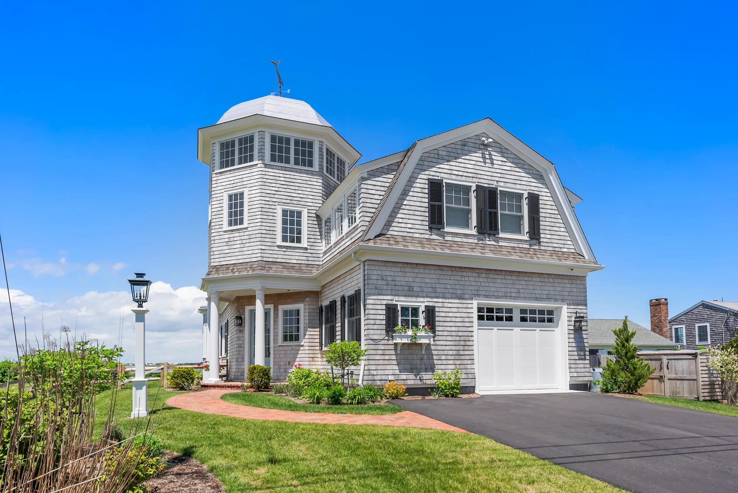 A large, gray, shingled house with multiple windows, black shutters, a white garage door, and a tower with a weathervane on top, set against a blue sky with some clouds.