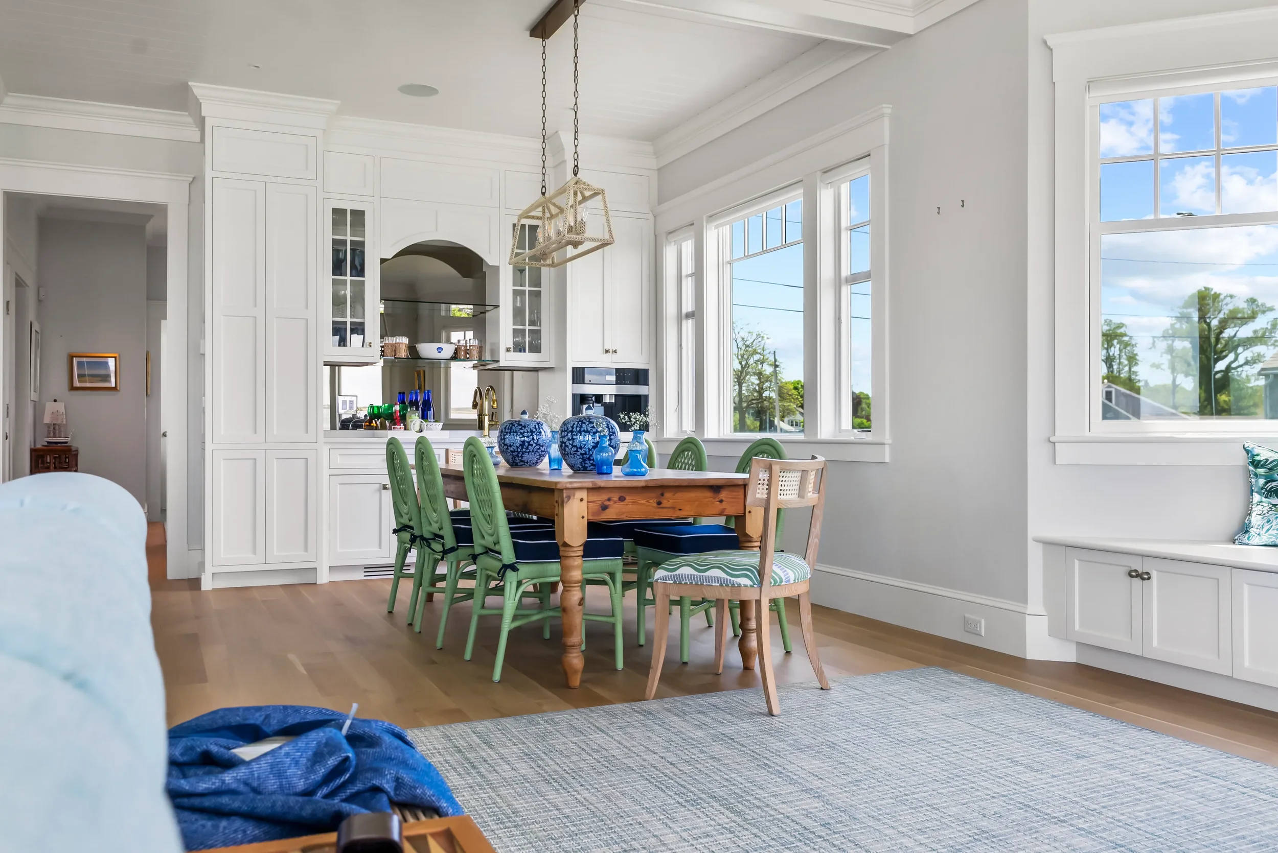 Bright dining room with white walls, large windows, and a wooden table surrounded by green chairs, decorated with blue vases, with a view of a neighborhood and trees outside.