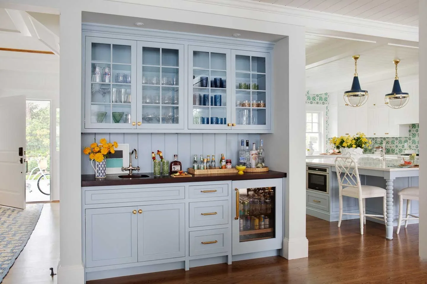 A kitchen area with light blue cabinetry and a dark wood countertop, decorated with a vase of yellow flowers and various liquor bottles. There is a small sink under the glass-fronted cabinets filled with glassware, and an adjacent bar area in another