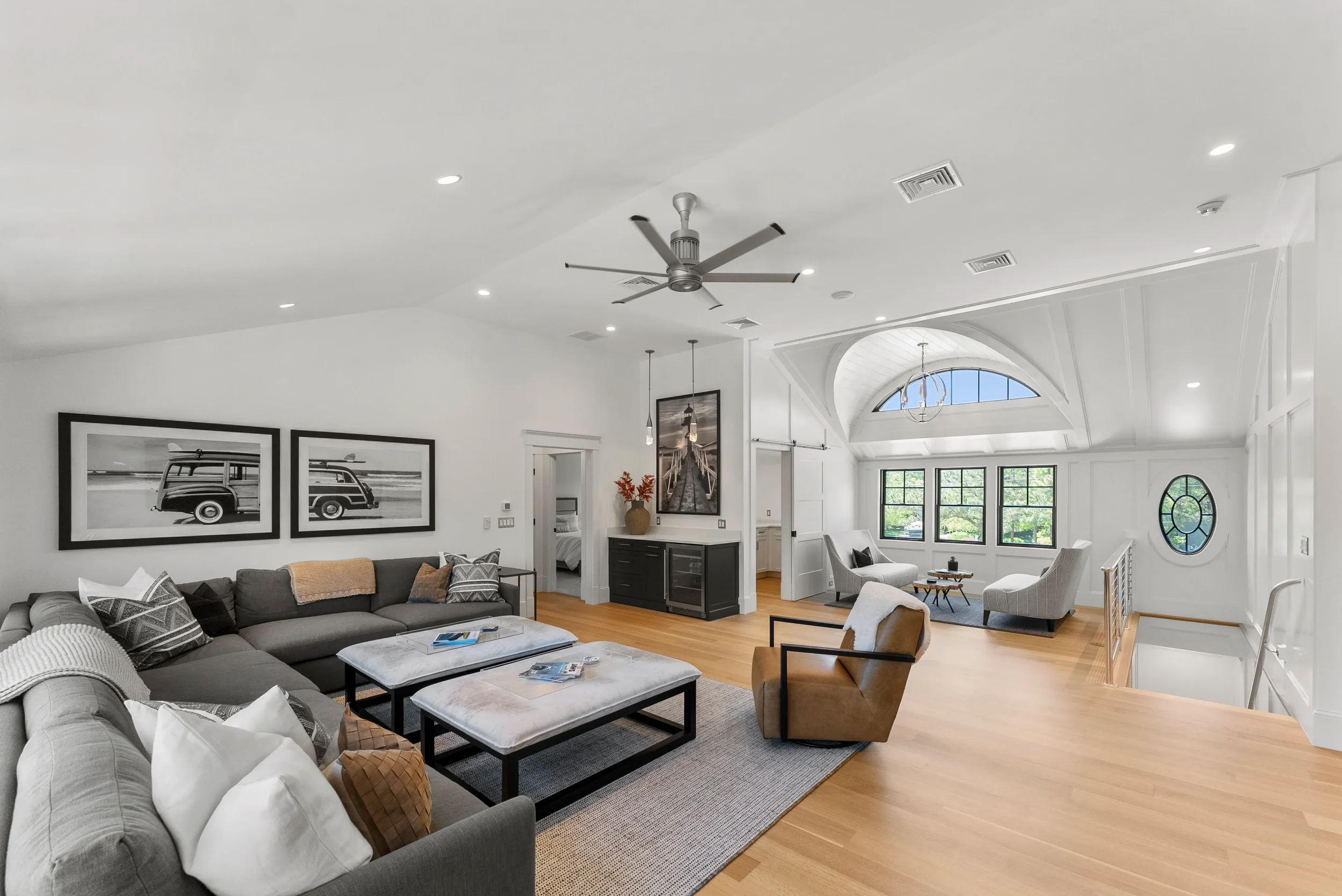 Spacious living room with white walls, hardwood floors, a large gray sectional sofa with pillows, two coffee tables, a tan armchair, and a white armchair near windows. Art photos of a vintage van on the wall, a black cabinet with a vase of orange flo