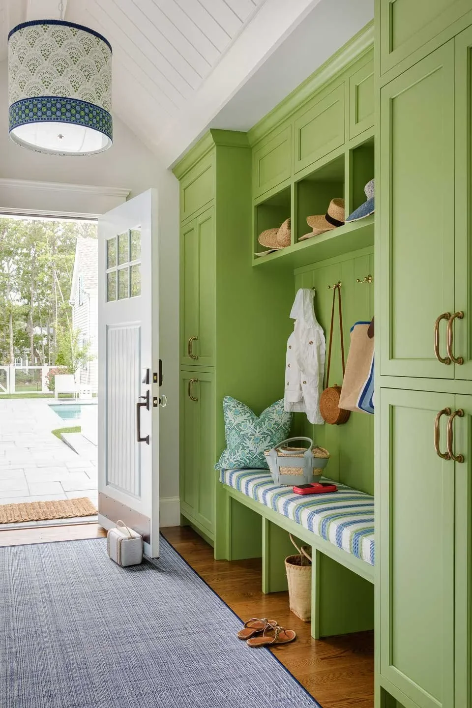 This image showcases a thoughtfully designed mudroom or entryway that blends functionality with coastal charm. The space features built-in green cabinetry with multiple compartments, hooks, and shelves—perfect for organizing everyday essentials like