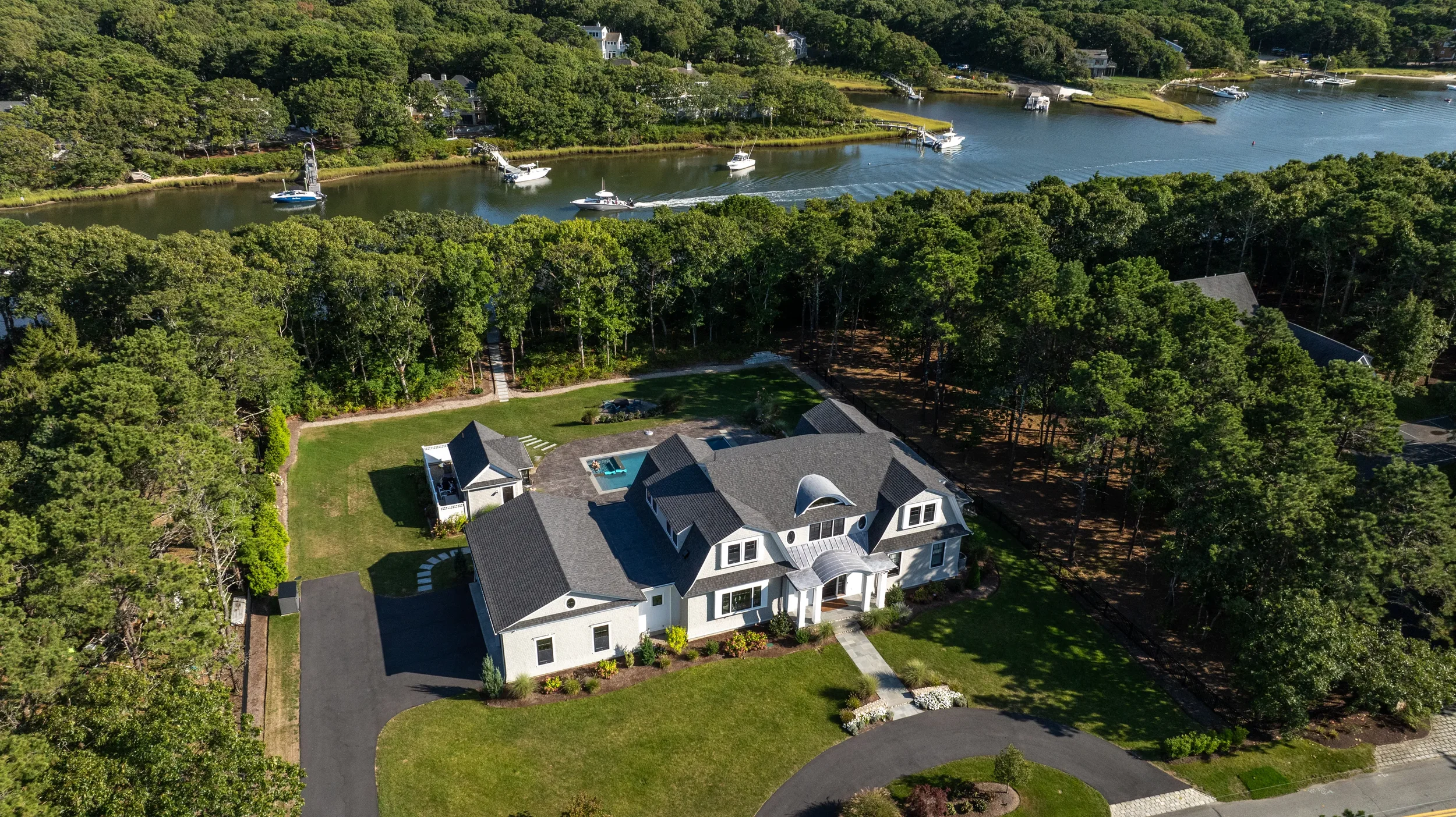 A large house with a gray roof, surrounded by a well-maintained lawn, trees, and landscaping. The backyard features a small pool. The house is near water, with boats visible on the adjacent river or creek.