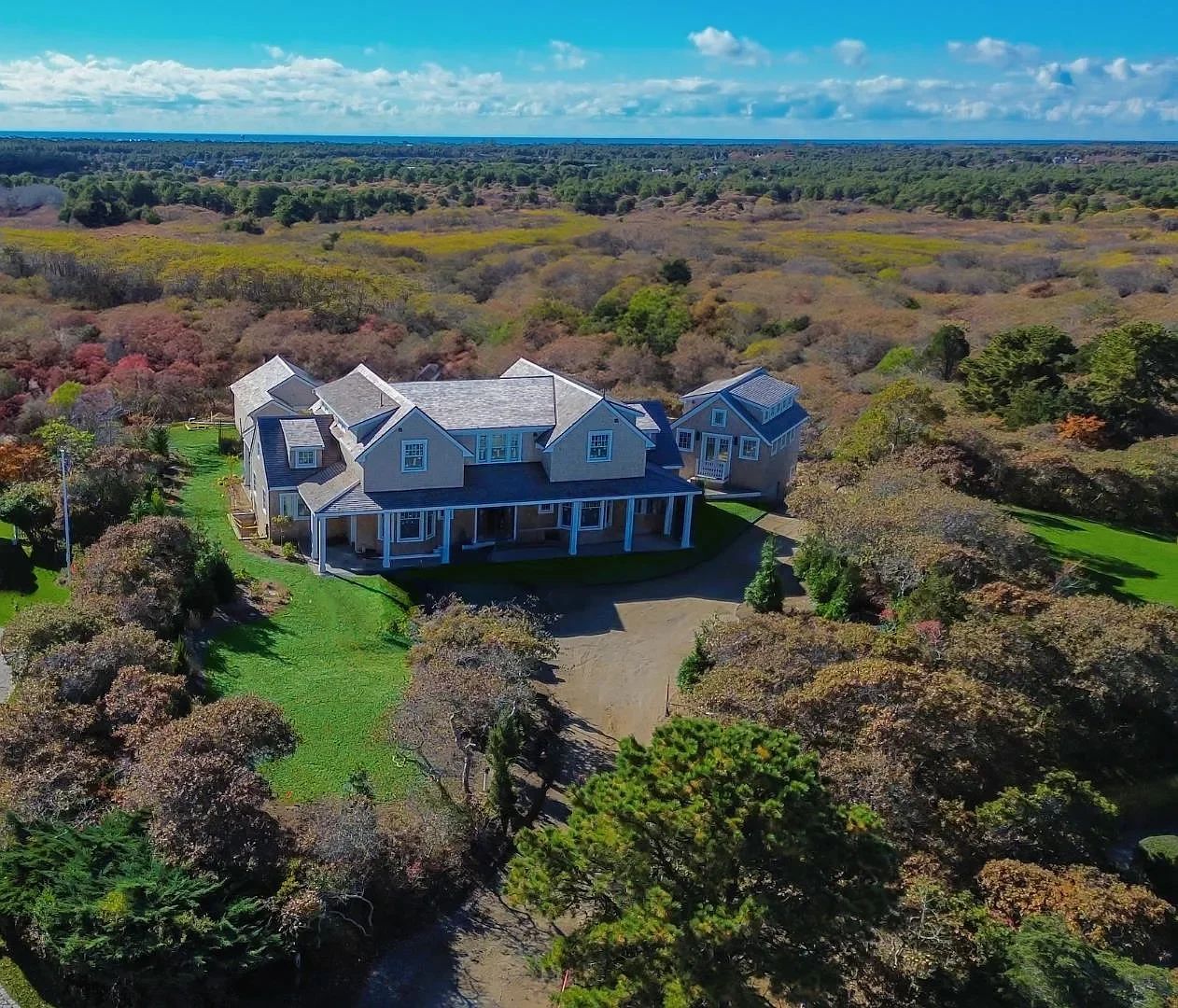 An aerial view of a large gray house with a spacious porch, surrounded by lush green trees and a landscape of rolling hills and fields in the background.