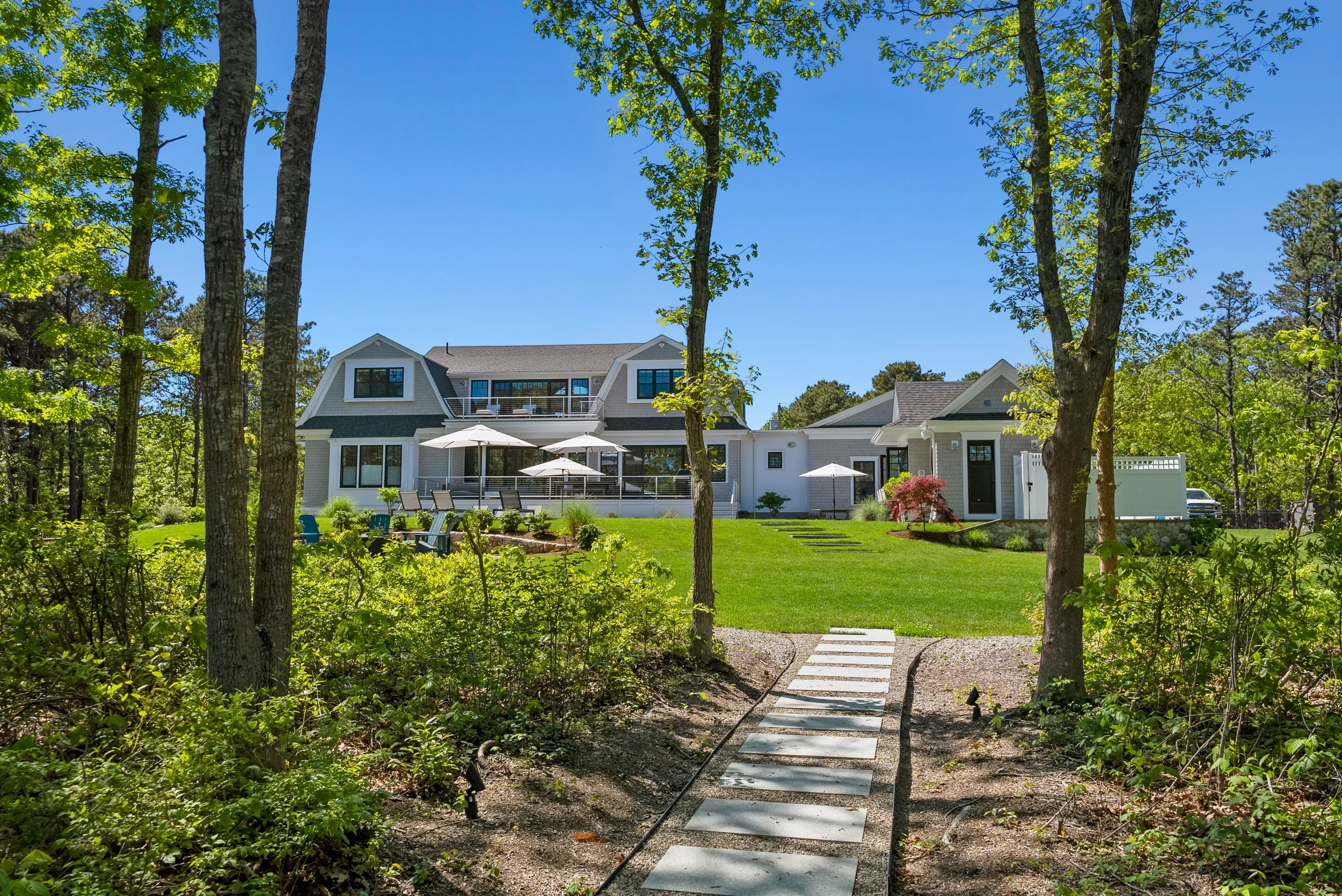 A large, modern house with a backyard patio features umbrellas, lounge chairs, and a well-maintained garden, surrounded by trees and lush green lawn under a clear blue sky.