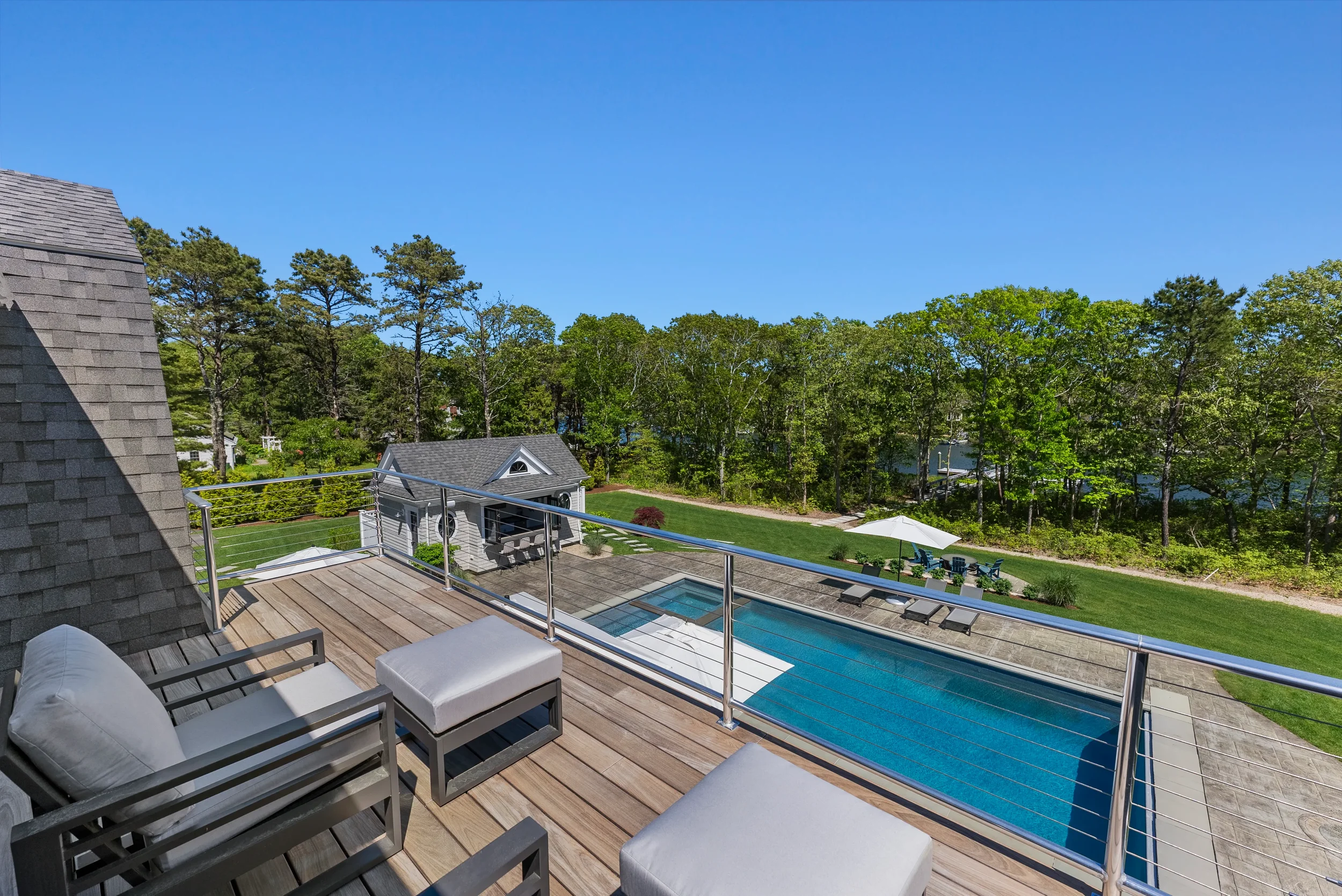 A view from a wooden balcony overlooking a backyard with a swimming pool, a patio with chairs and an umbrella, a shed, and green trees under a bright blue sky.
