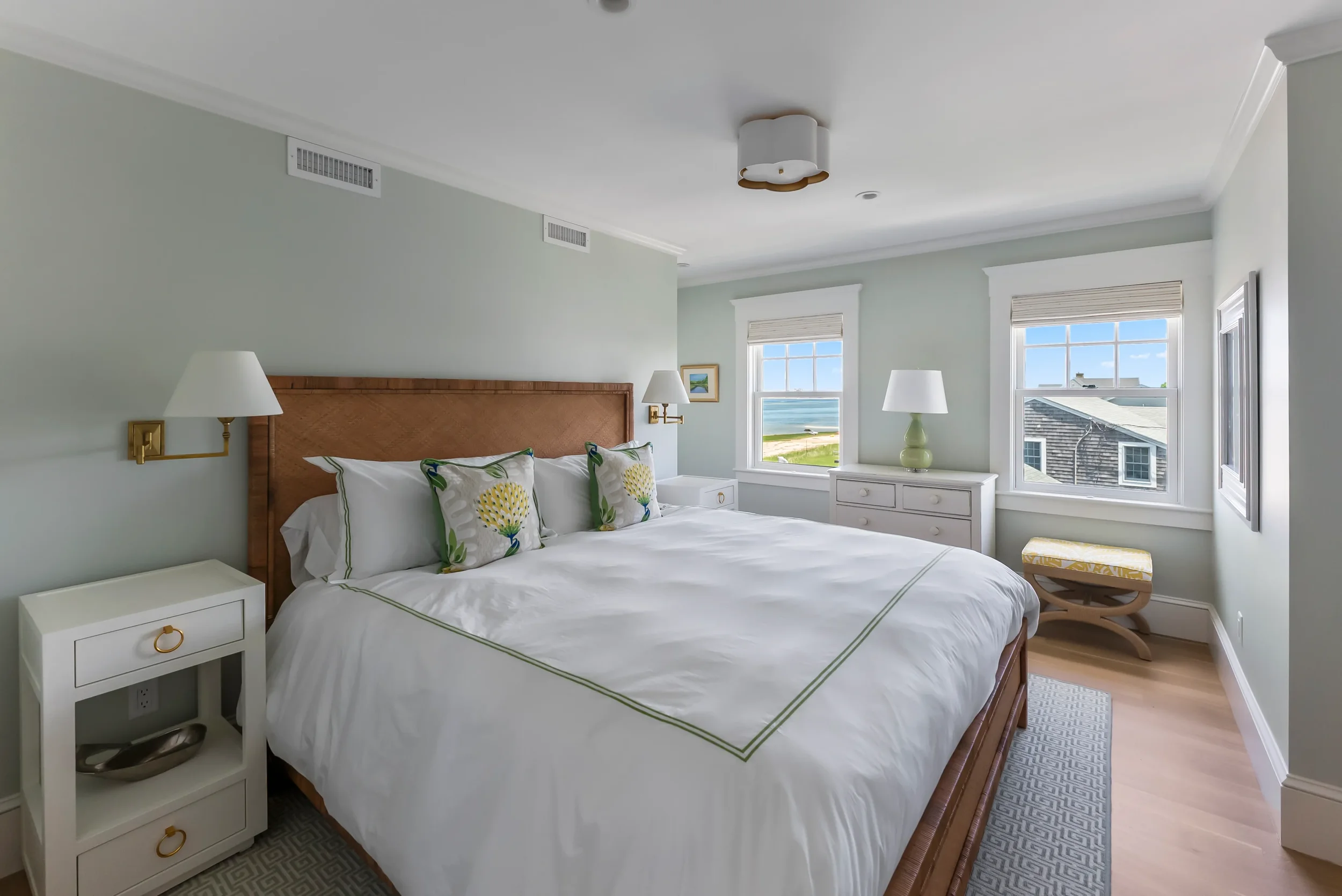 Bright hotel bedroom with a large bed, white bedding, and decorative pillows. Two bedside tables with lamps, a chest of drawers, and two windows showing a beach view. Light-colored walls and hardwood floors.
