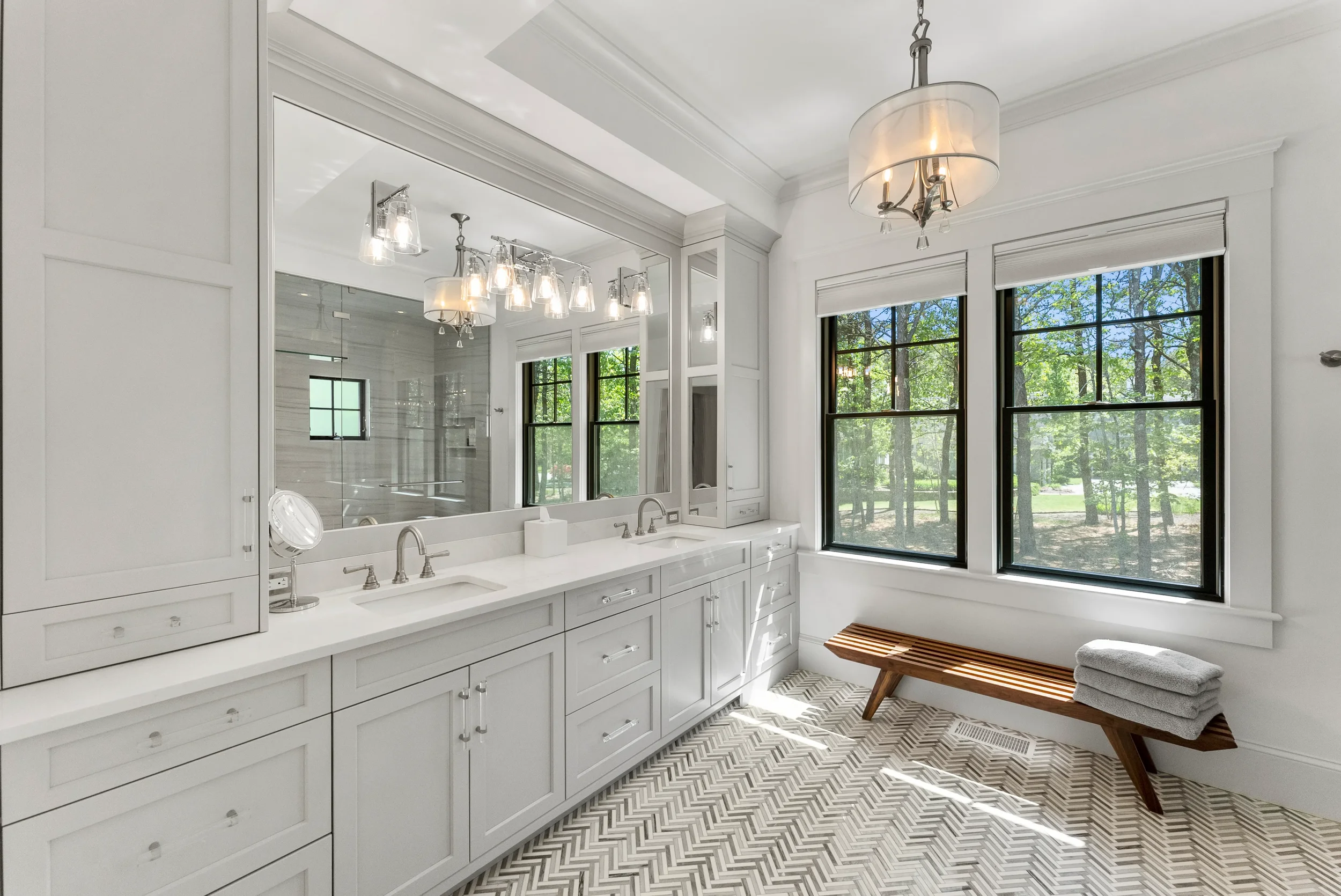 A bright bathroom with white cabinetry, two sinks, a large mirror, and black-framed windows showing greenery outside. A wooden bench with folded towels is beneath the windows, and there's a ceiling light fixture.