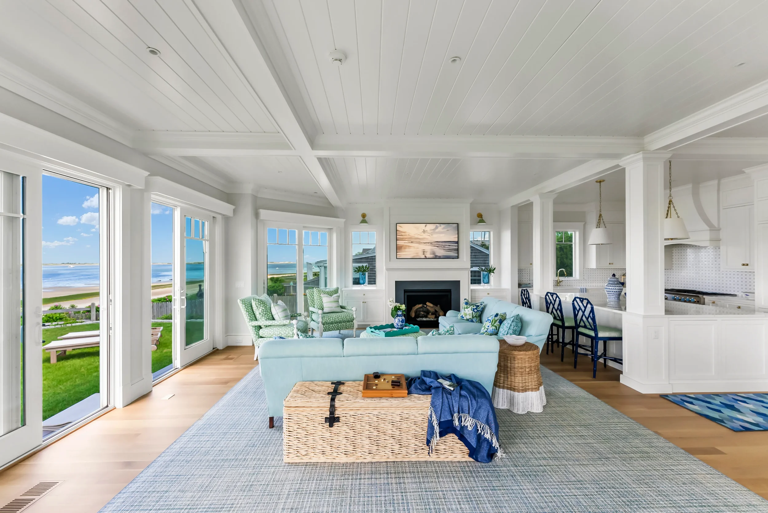 Bright living room with large windows overlooking a beach, white walls, white ceiling, light hardwood floor, blue sofa, green armchairs, fireplace, nautical decor, and a view of the ocean.