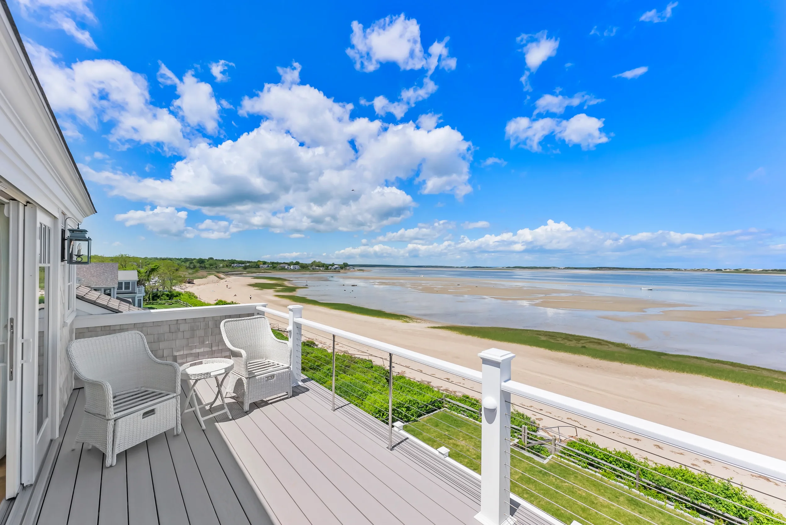A spacious balcony overlooking a beach with sandy shore, water, and scattered clouds in a blue sky.